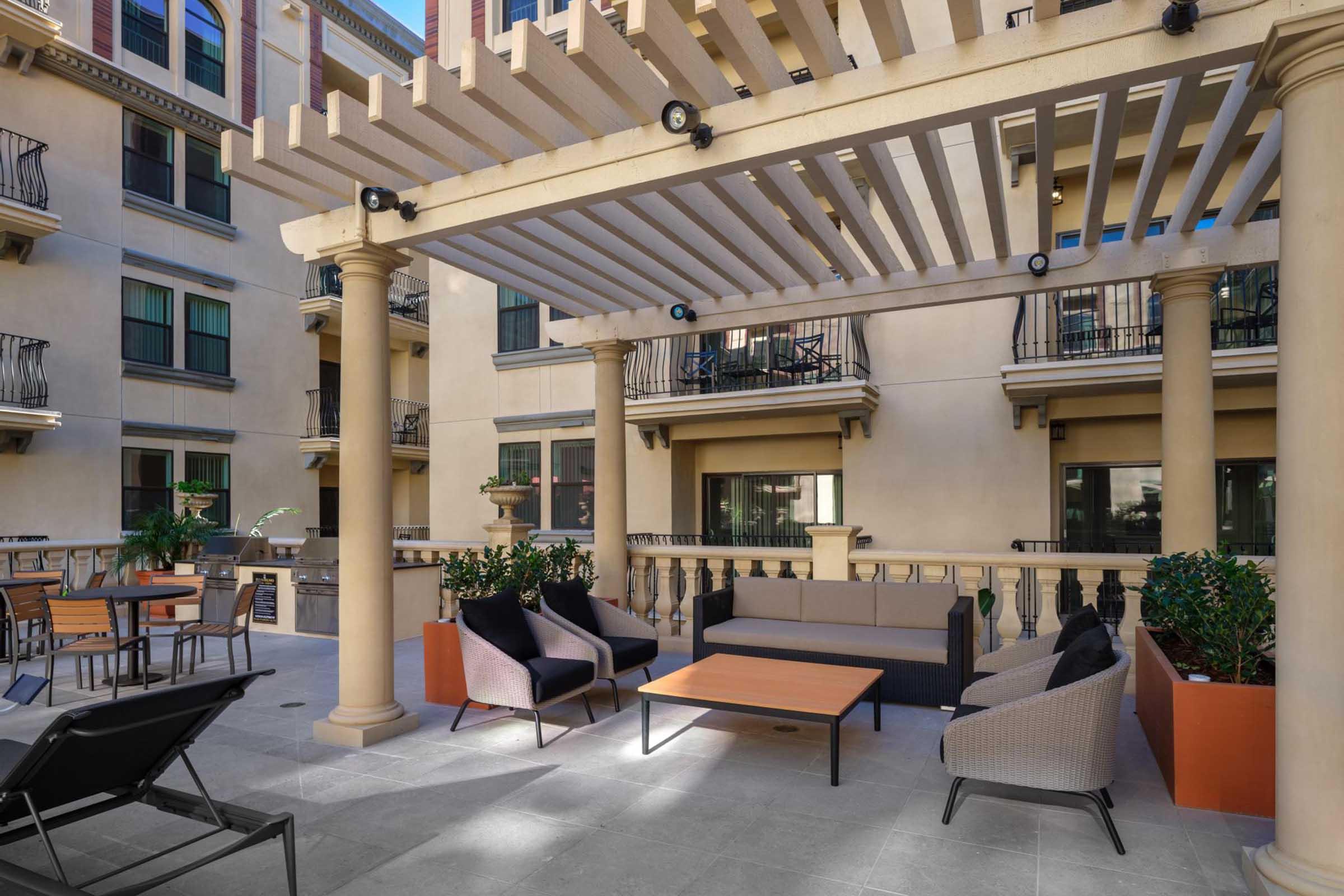 A cozy outdoor lounge area featuring a pergola, comfortable seating, and a coffee table. Surrounded by potted plants, it is located within a well-designed building with balconies. Sunlight filters through the slats of the pergola, creating a welcoming atmosphere for relaxation or socializing.