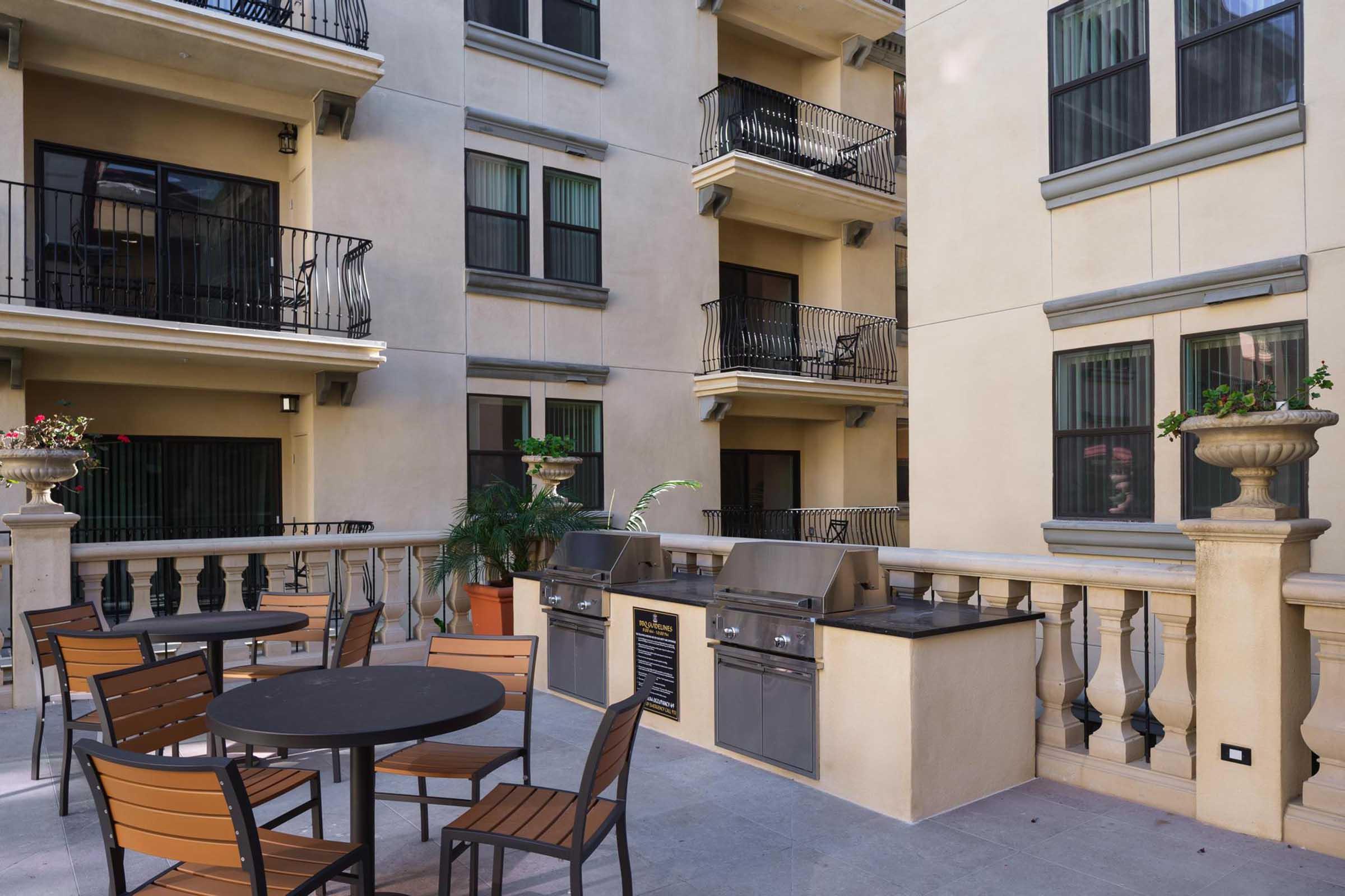 Outdoor lounge area featuring a large table with chairs, two barbecue grills, and decorative potted plants. The backdrop includes balconies of a multi-story building with wrought iron railings, creating a welcoming space for residents to gather and socialize.