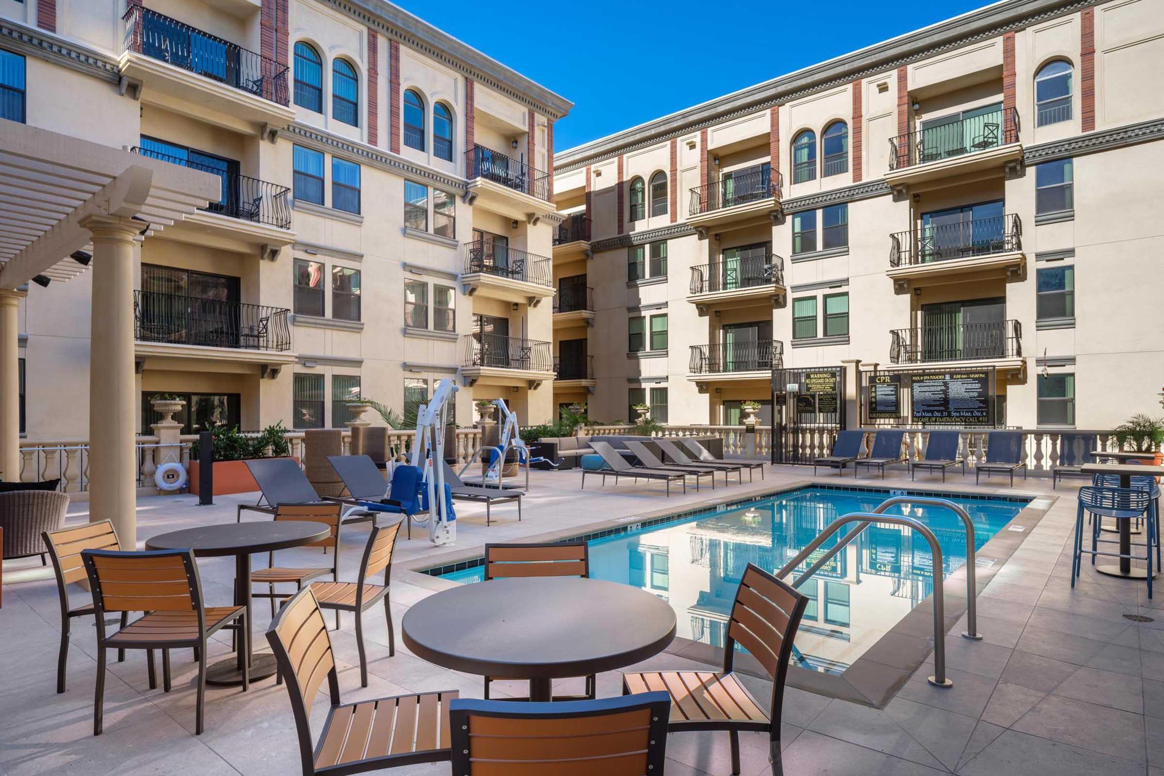 A tranquil pool area featuring lounge chairs and tables, surrounded by modern apartment buildings. Sunlight reflects off the water, with decorative elements like potted plants enhancing the inviting atmosphere. Ideal for relaxation and leisure.