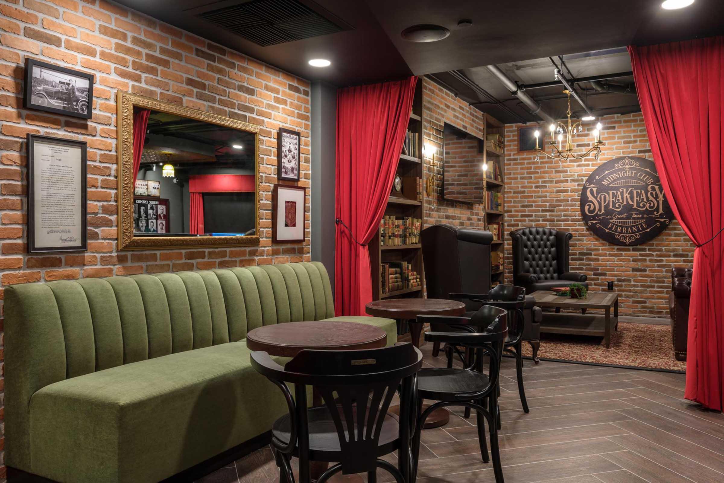 Cozy interior of a speakeasy-style lounge featuring exposed brick walls, a green velvet sofa, wooden tables, and black chairs. A large mirror reflects the ambience, with red curtains and dim lighting adding to the relaxed atmosphere. A sign on the wall reads "Speakeasy," contributing to the vintage aesthetic.