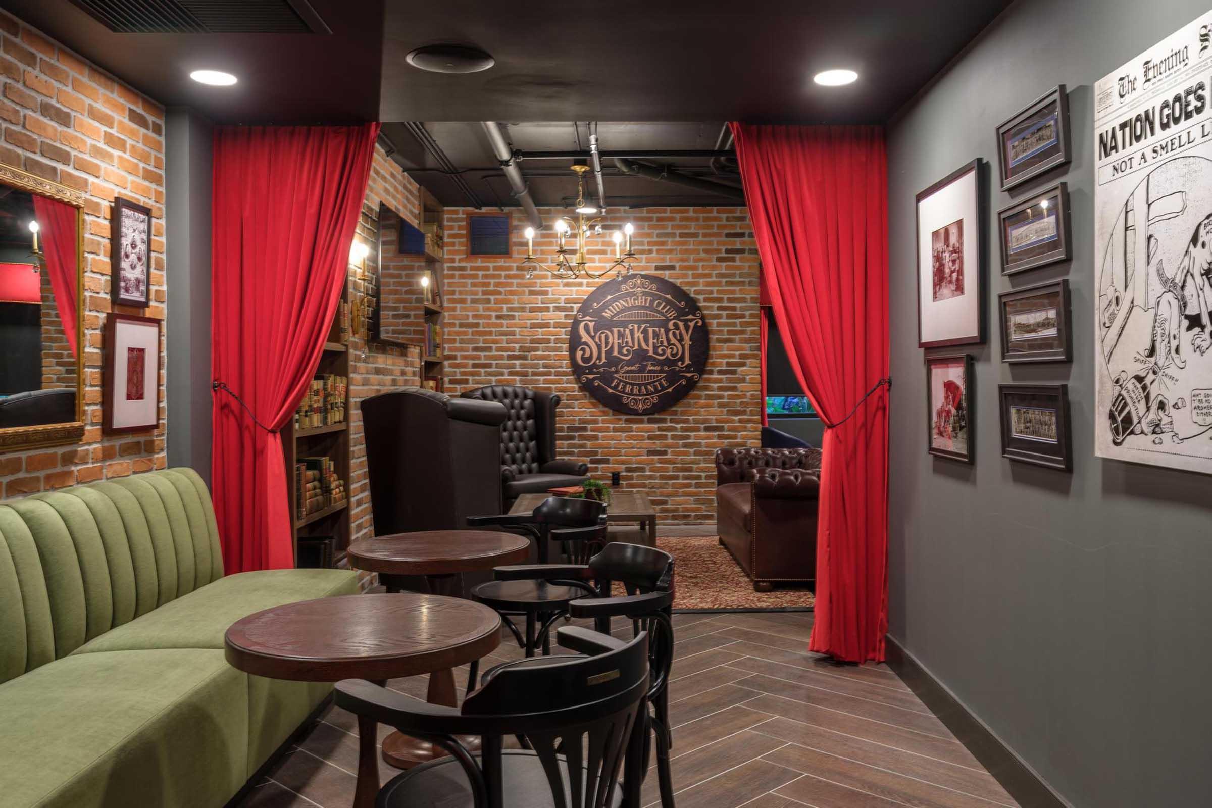 A cozy speakeasy-style lounge featuring exposed brick walls, plush seating, and warm lighting. A green velvet sofa, wooden tables, and stylish decor invite relaxation. Red curtains close off an intimate space, adorned with vintage posters and a playful logo, creating a charming and inviting atmosphere.
