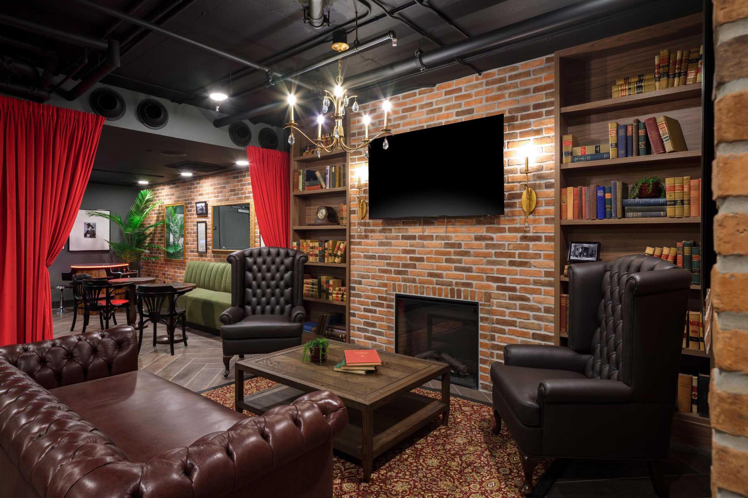 Cozy lounge area featuring a brick wall, dark wood bookshelves filled with books, two leather armchairs, a coffee table with decorative items, a fireplace, and a large television mounted on the wall. Red curtains frame the space, adding warmth to the atmosphere. A green upholstered sofa is visible in the background.