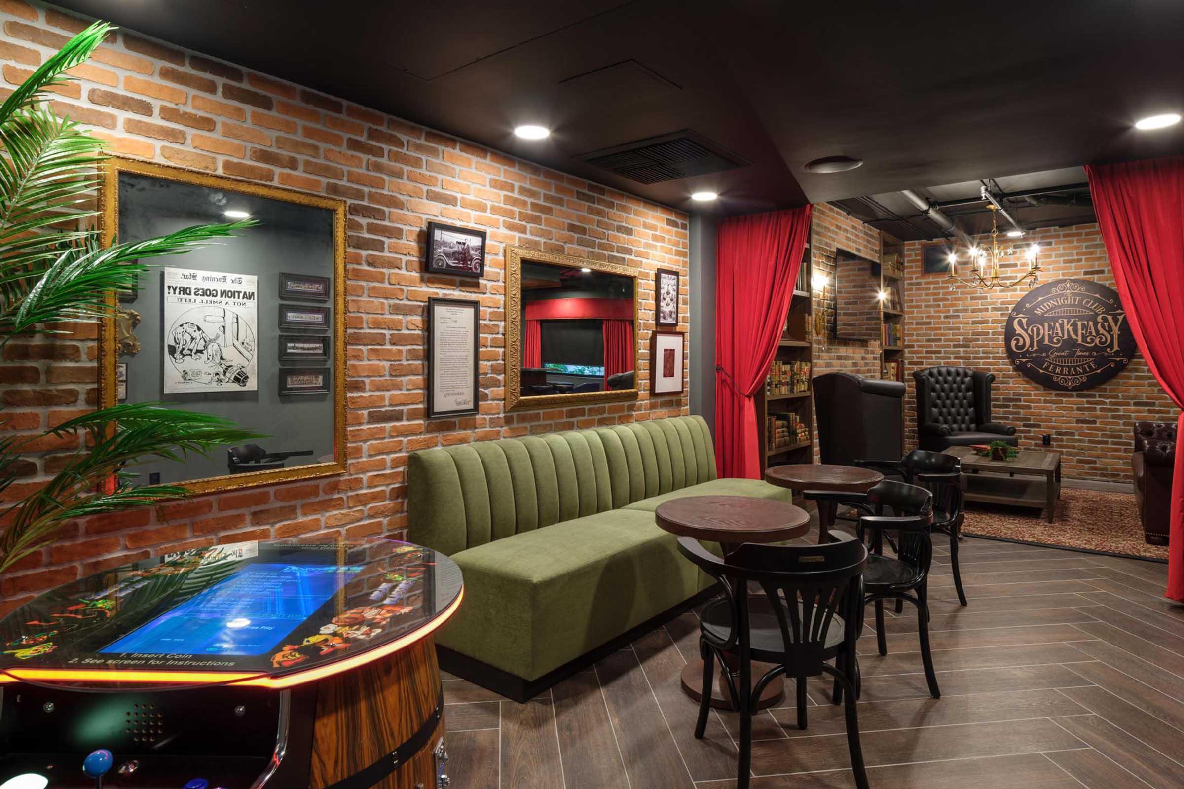 A stylish speakeasy-themed lounge with exposed brick walls, featuring a green velvet sofa, small round tables, and black leather armchairs. Red curtains frame the entrance, and a vintage pinball machine adds a playful touch to the cozy, elegant atmosphere. Subtle lighting enhances the inviting ambiance.