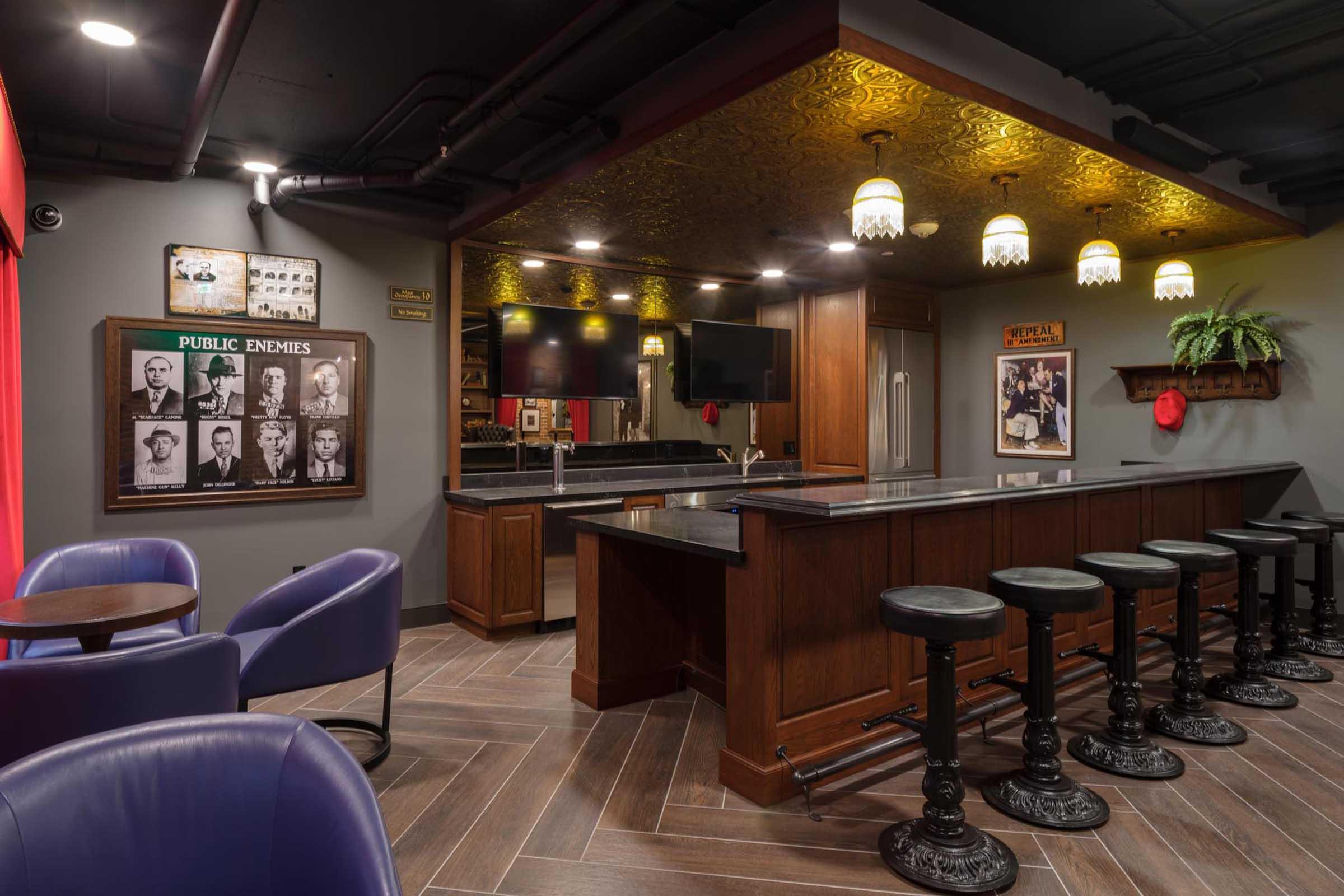 A stylish home bar featuring dark wood cabinets, a black countertop, and high-backed stools. The walls are adorned with framed historical photos and decorative elements, while warm lighting fixtures provide an inviting ambiance. Cozy seating is arranged nearby, enhancing the bar's social atmosphere.