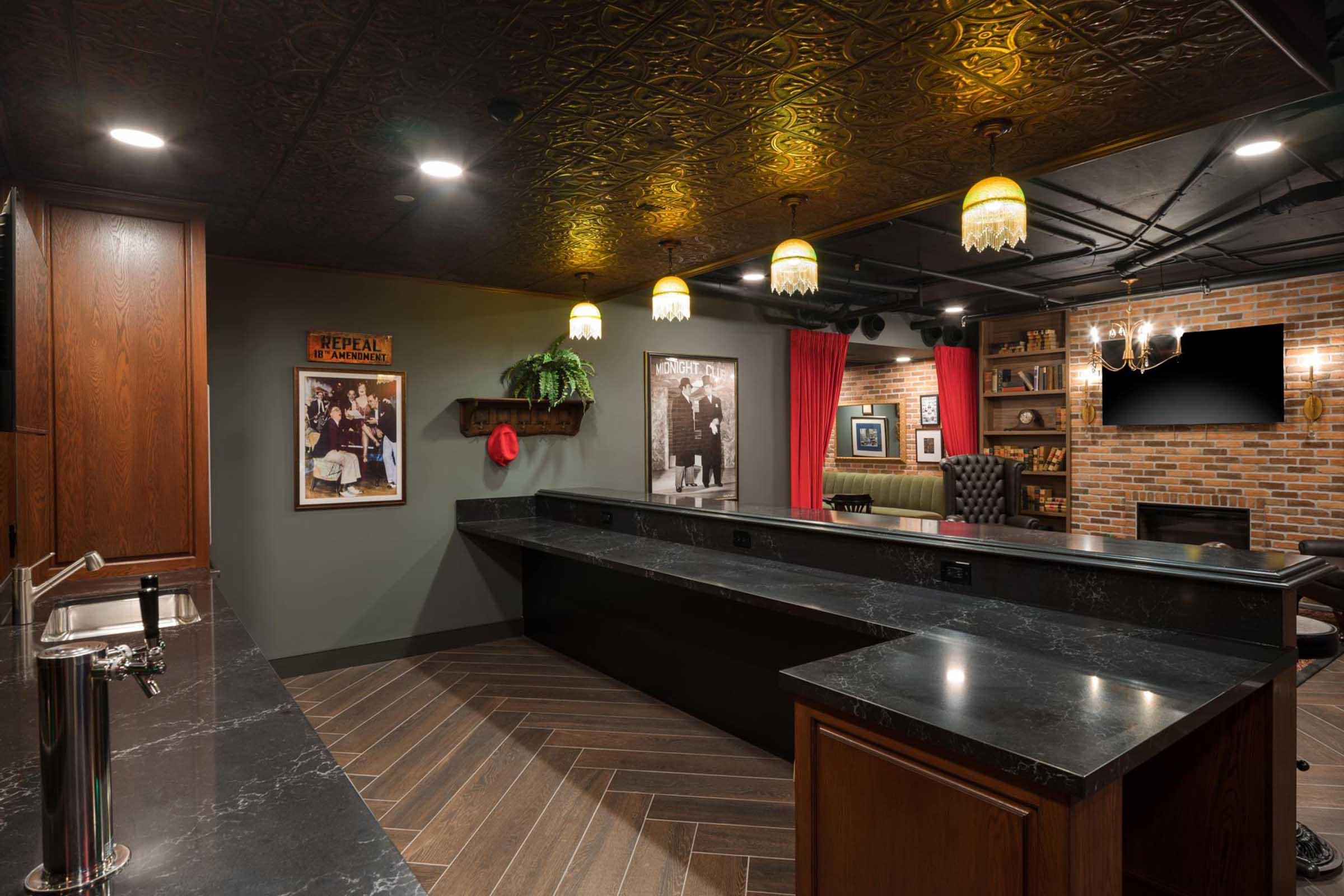 A stylish bar area featuring a dark countertop, vintage decor, framed black-and-white photos, and warm pendant lighting. The space includes a sink, wooden cabinetry, and a backdrop of red curtains and exposed brick, creating a cozy and inviting atmosphere for social gatherings.