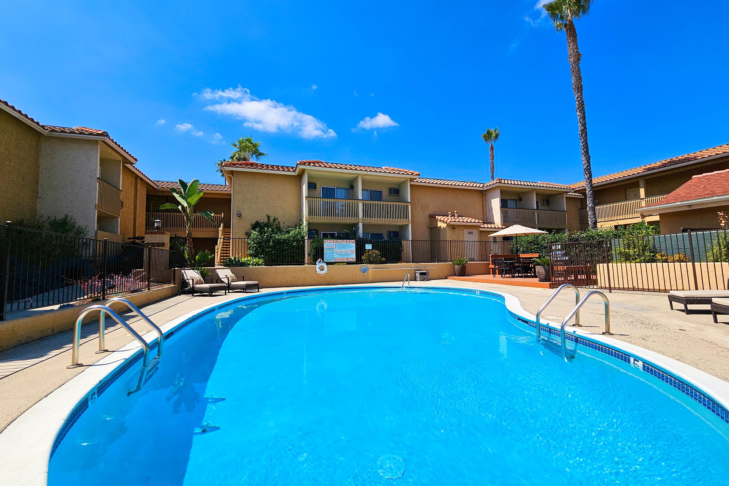 A bright blue swimming pool surrounded by lounge chairs and palm trees, set against a backdrop of a multi-story building with balconies. The clear blue sky adds to the inviting atmosphere of the pool area.