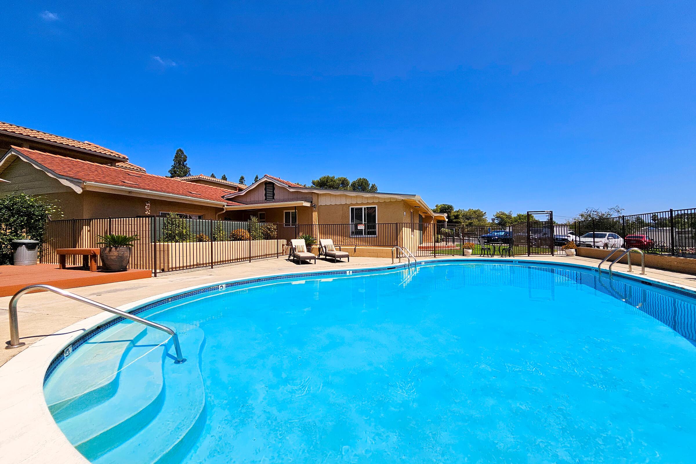 A clear blue swimming pool surrounded by a safety fence, with patio chairs nearby. In the background, a residential building with a sunny blue sky overhead and trees visible. The scene conveys a relaxed outdoor setting, ideal for leisure or recreation.