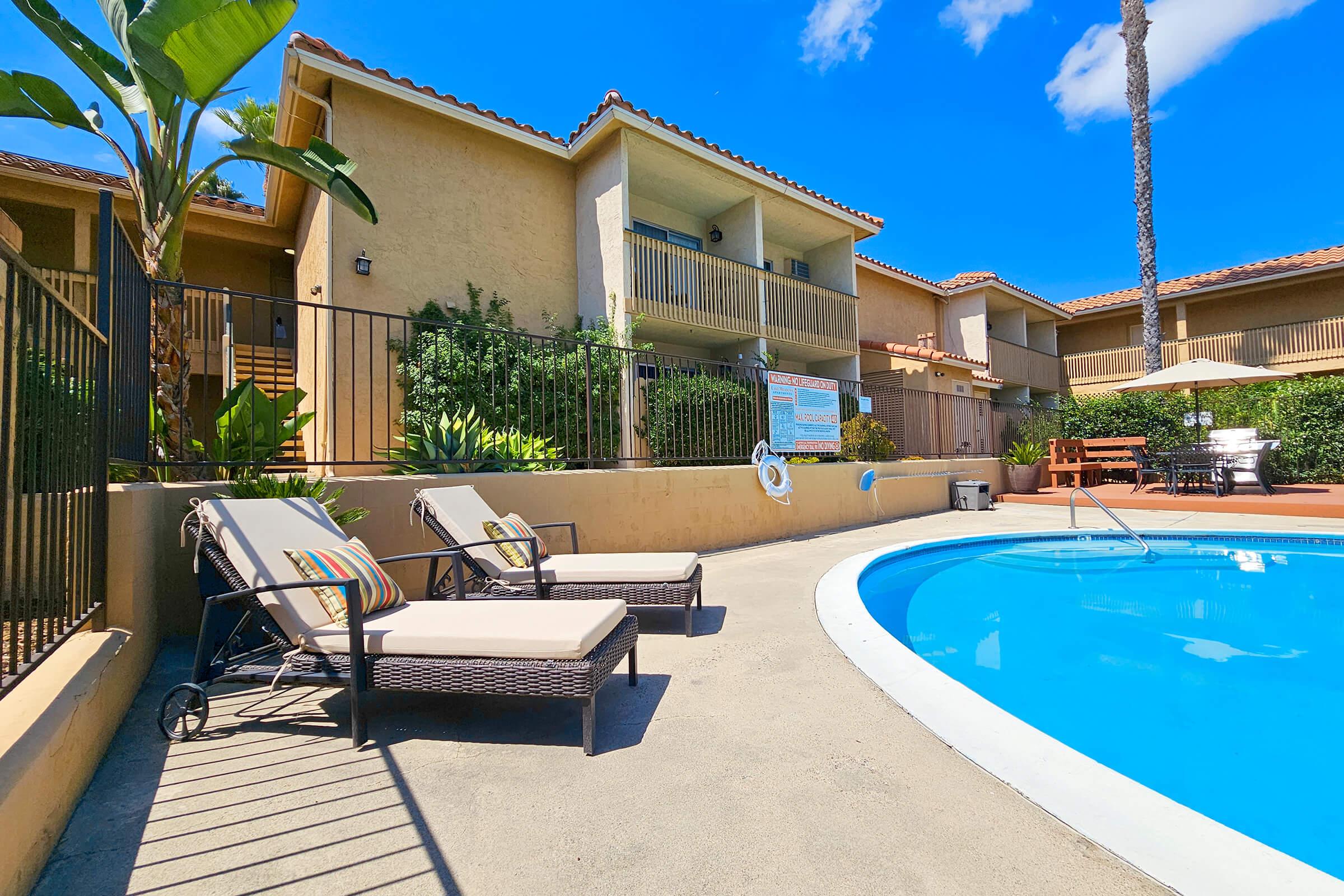 A clear blue swimming pool in a residential area with lounge chairs placed beside it. The background features two-story buildings with balconies, surrounded by palm trees and greenery. A sign and a seating area are visible near the pool, all under a bright blue sky.
