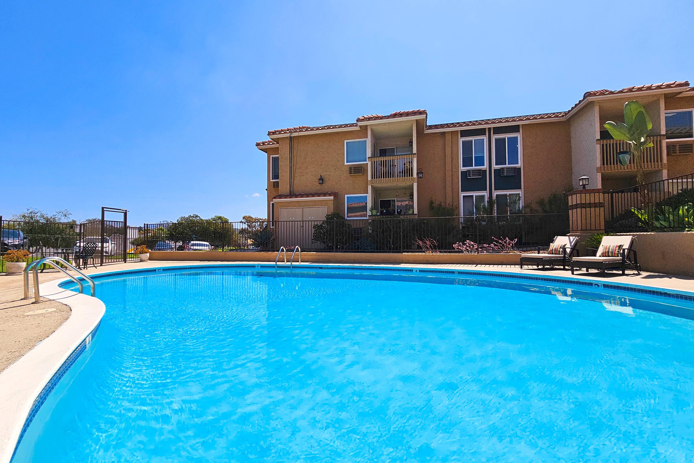 A clear blue swimming pool in the foreground, surrounded by a tan concrete deck. In the background, there is a multi-story residential building with balconies. Lush greenery and a few lounge chairs are visible near the pool area, all under a bright blue sky.