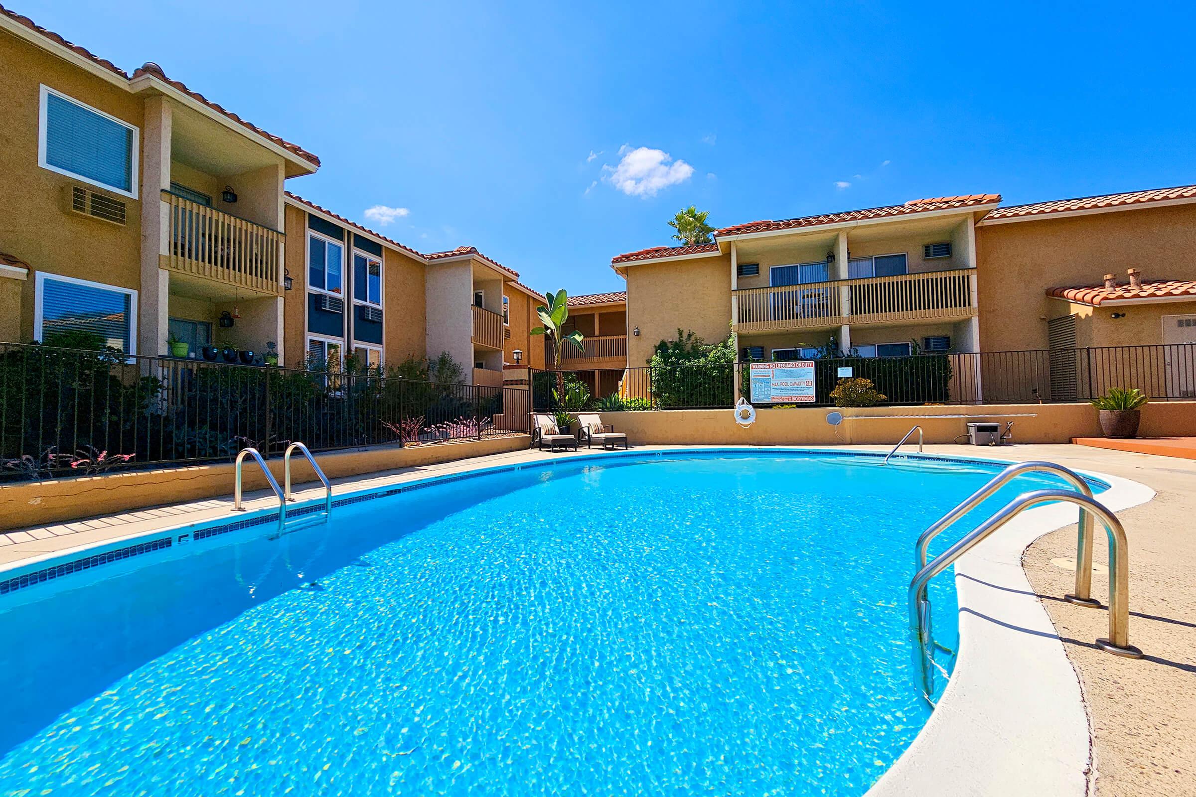 A crystal-clear swimming pool surrounded by beige apartment buildings. Lush greenery and potted plants add to the landscape. The sky is bright blue with a few white clouds, creating a sunny and inviting atmosphere. Sun loungers sit near the poolside, perfect for relaxation.