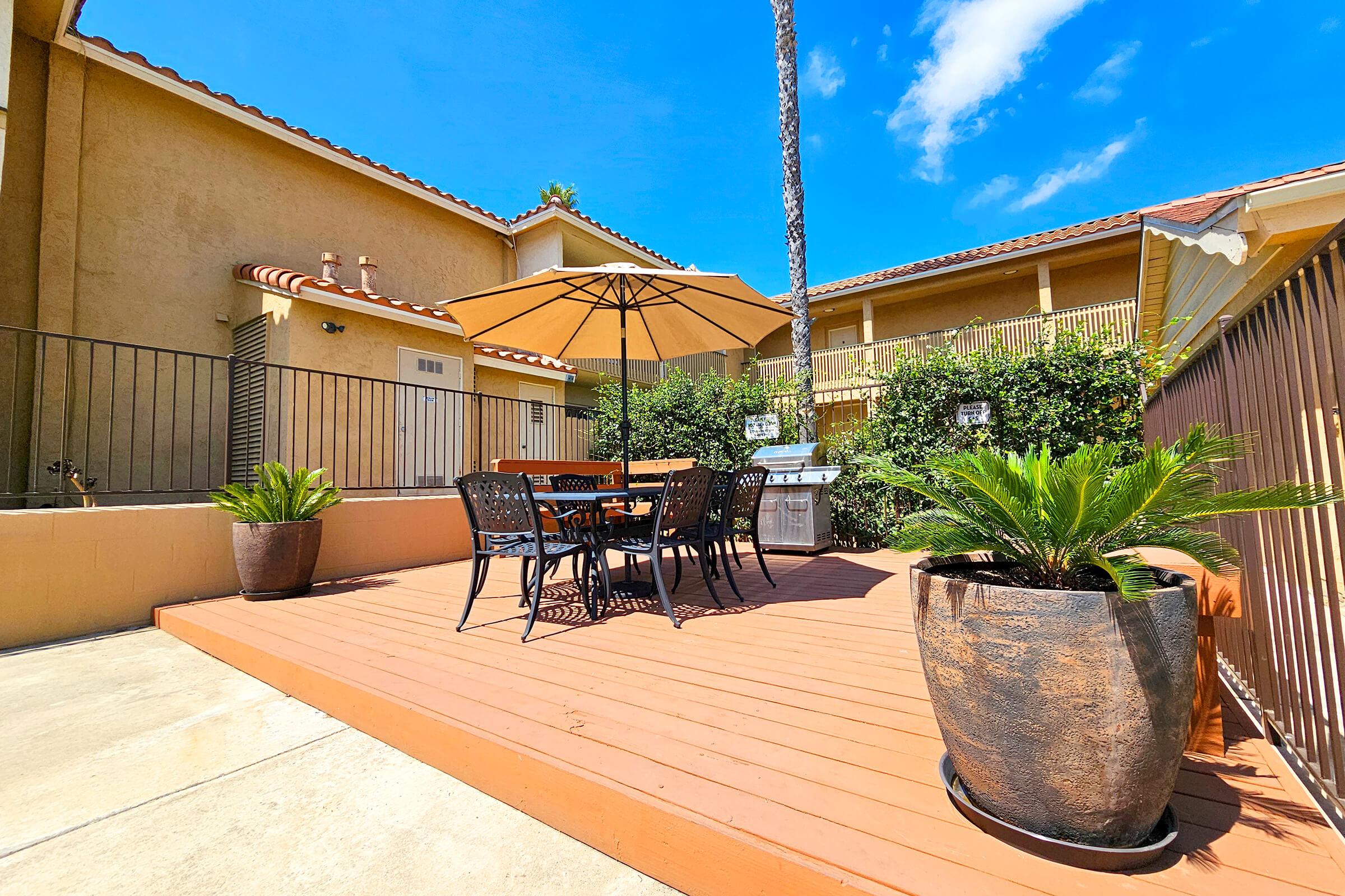 A sunny outdoor patio area featuring a wooden deck with a dining table and four chairs under a large umbrella. Two potted palm plants are on either side, and in the background, there are buildings with balconies. The sky is bright blue with a few fluffy clouds.