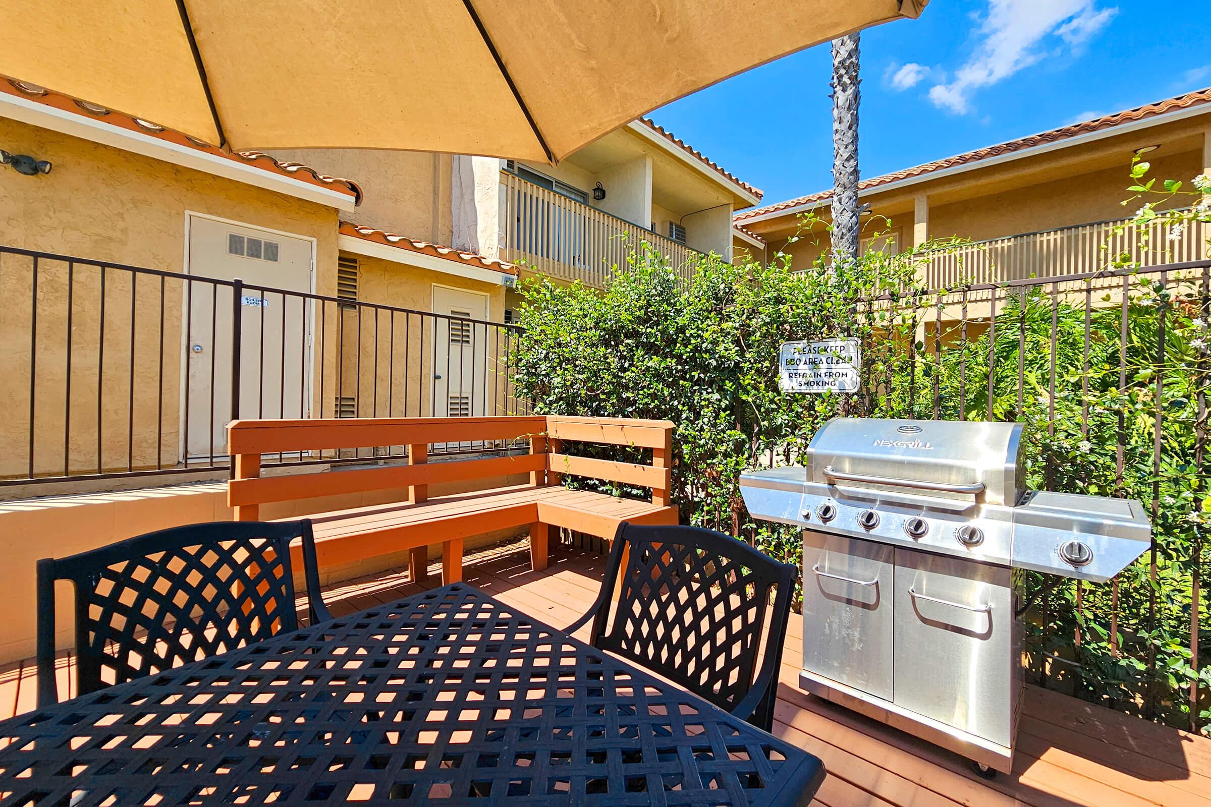 A patio area featuring a black outdoor table and chairs under a sun umbrella, with a stainless steel gas grill nearby. In the background, a two-story building with balconies and green foliage surrounds the space, creating a cozy outdoor setting. Bright blue sky visible above.