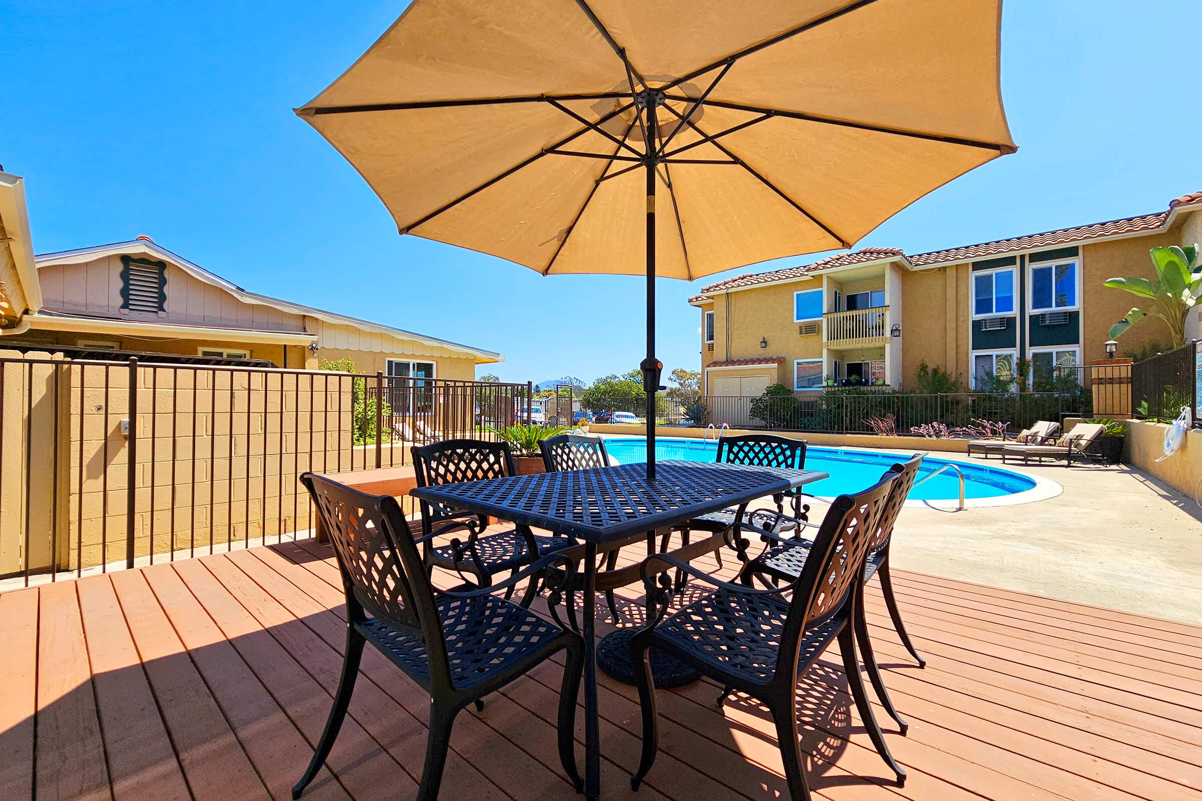 A sunny outdoor patio features a black metal dining table with matching chairs, shaded by a large umbrella. In the background, a blue swimming pool is visible, along with beige residential buildings and greenery, creating a relaxing atmosphere.