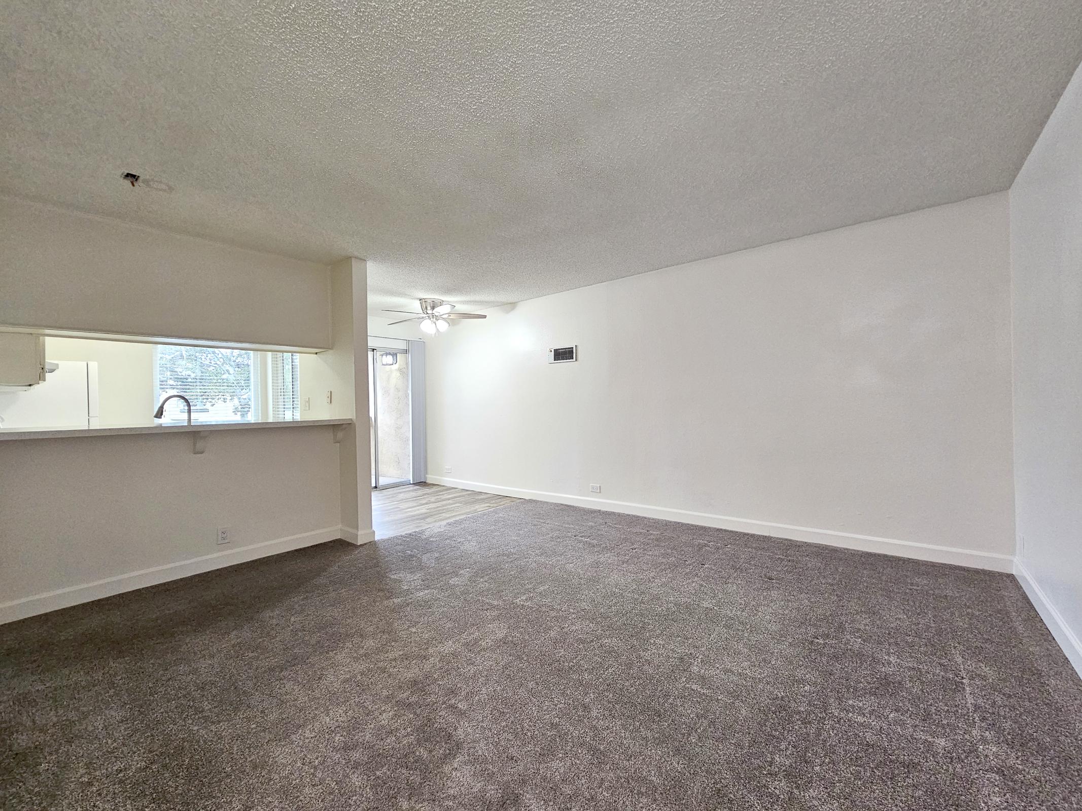 Empty living room with light-colored walls and dark carpet. A ceiling fan is visible, and there is a doorway leading outside. An open kitchen area is seen in the background, with a bar counter separating it from the living space. The overall atmosphere is bright and spacious.