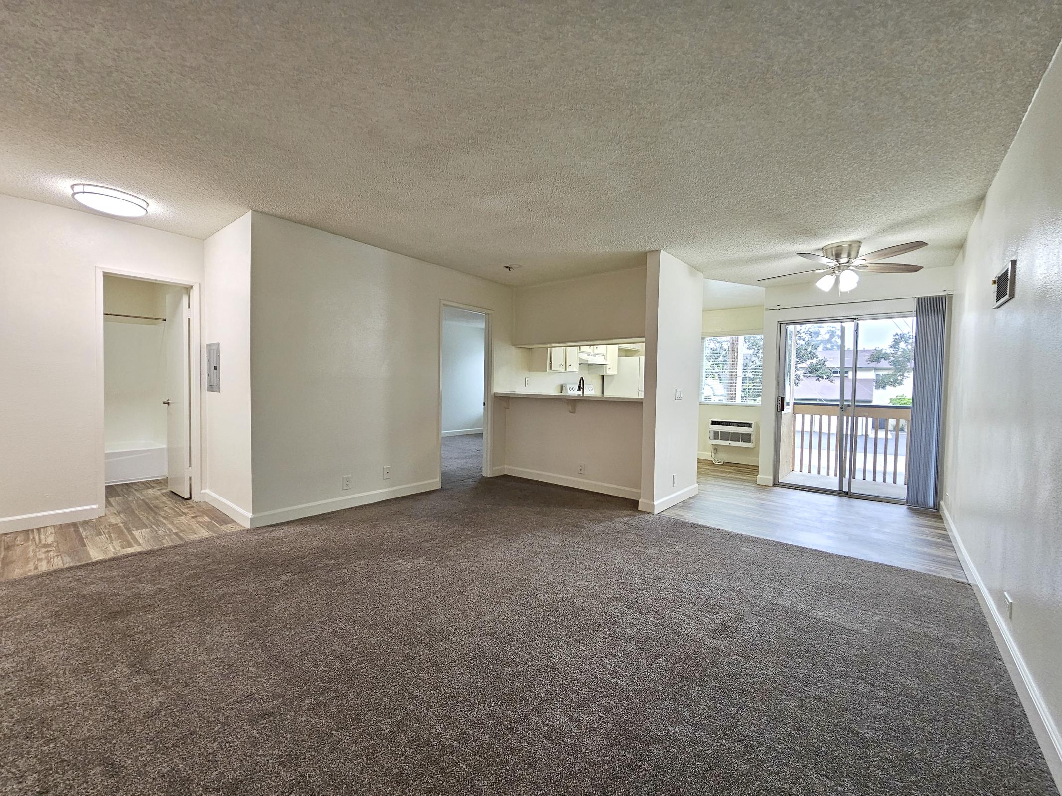 Spacious living area with carpet flooring, a ceiling fan, and large windows allowing natural light. The room features an open layout leading to a kitchen with a bar. A sliding glass door provides access to a balcony, and there's an adjoining room that appears to be a closet.