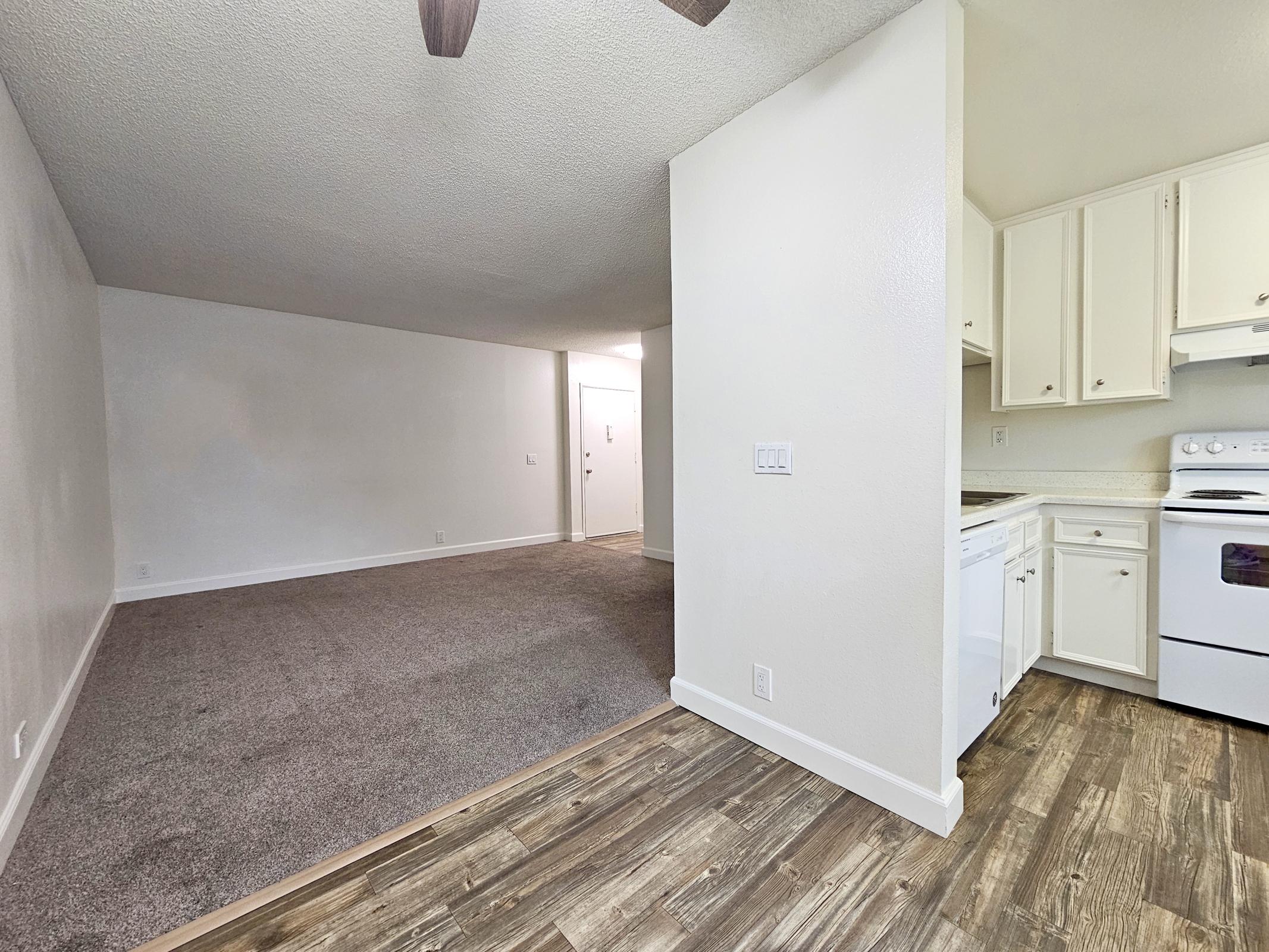 An interior view of an apartment featuring a spacious living area with beige carpet, a ceiling fan, and an open kitchen with white appliances and cabinets. The kitchen is partially visible, showing the stove and countertop, while the living area is well-lit with a light-colored wall.