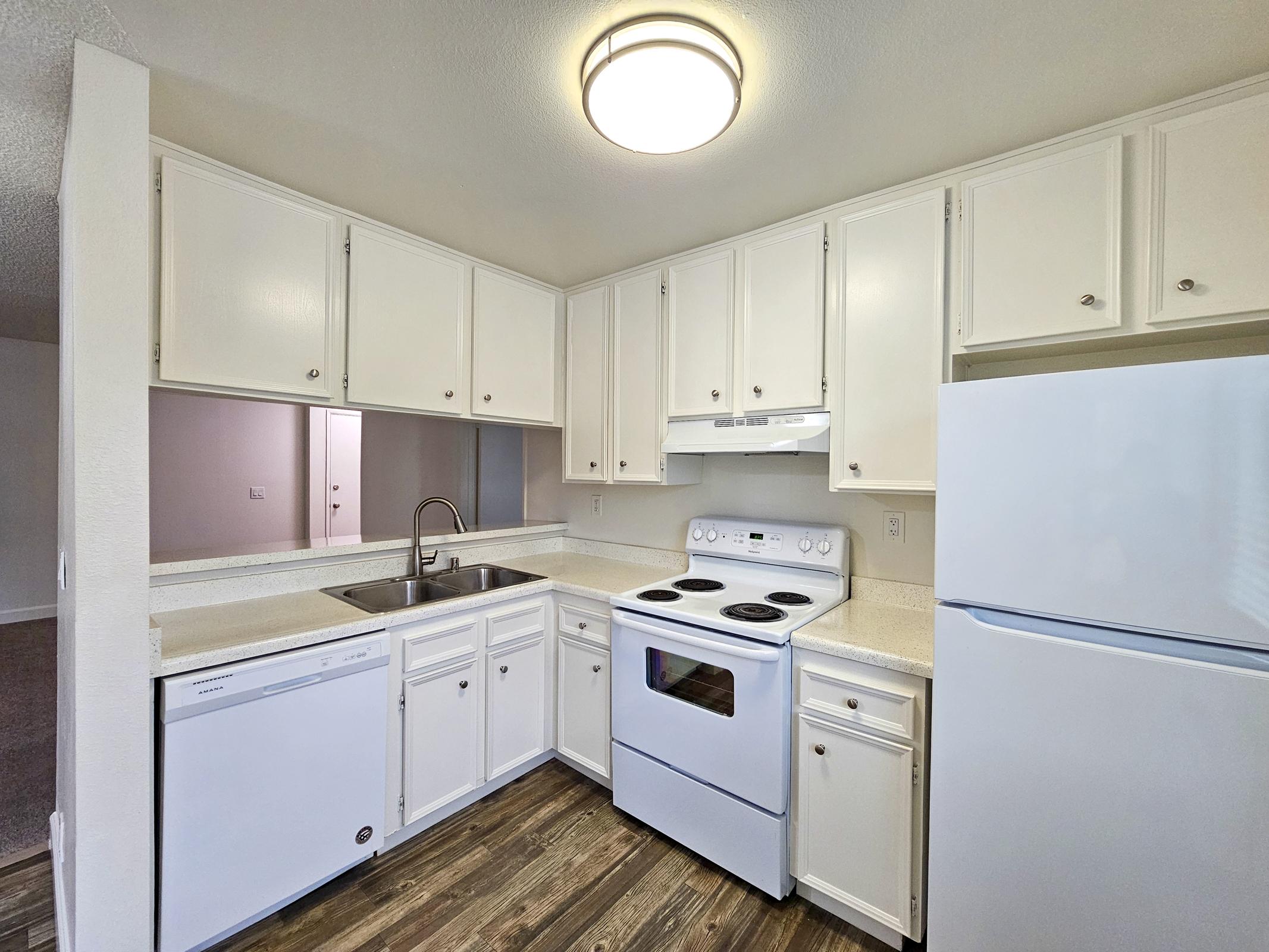 A modern kitchen with white cabinets, a sink, a stove, and a refrigerator. The countertop is light-colored, and there is a dishwasher below the countertop. The kitchen features overhead lighting and a wood-like floor. The background shows a doorway leading to another room.