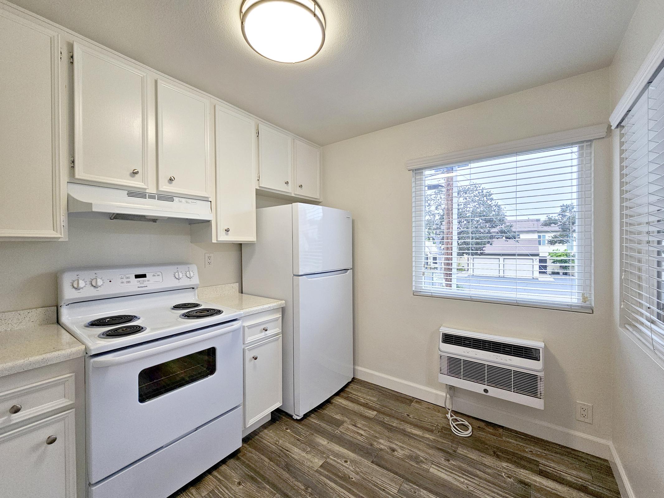 A bright kitchen featuring white cabinets, a white refrigerator, and a white stove with an oven. Natural light streams in through a window with blinds, and there is an air conditioning unit on the floor. The flooring is a light wood design, creating a clean and modern look.