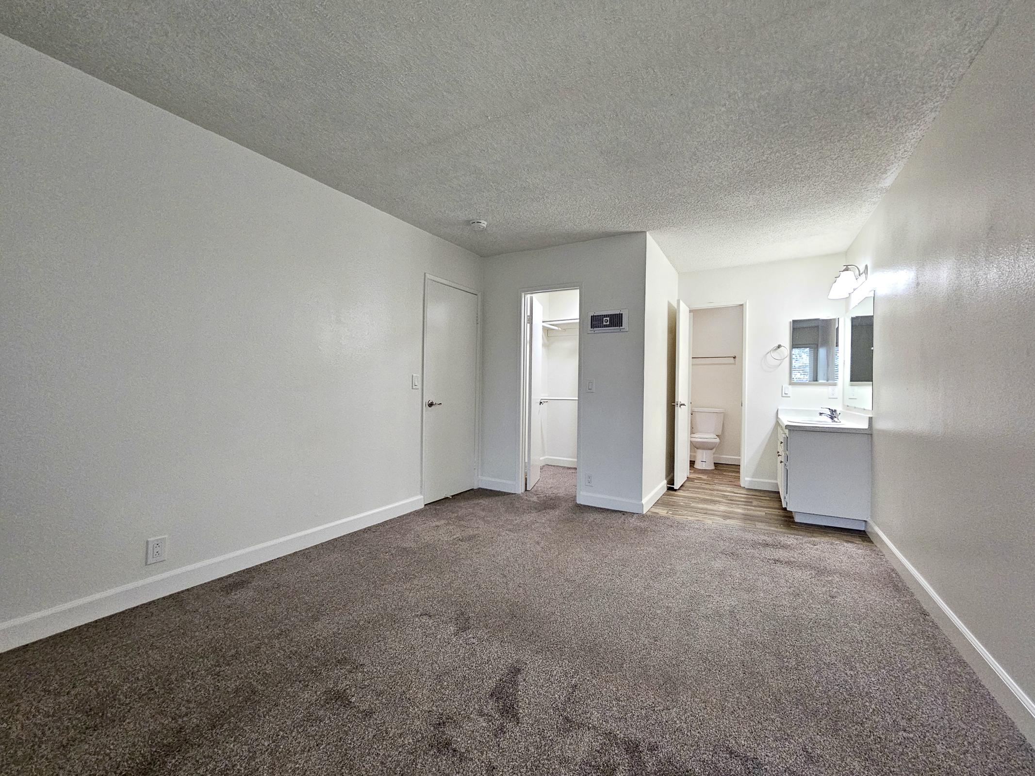 A vacant room featuring beige carpeting and white walls. To the right, a bathroom door is visible, and a vanity area with a mirror and light fixture is next to it. The room has a spacious layout with a single door leading to another area, illuminated by natural light.