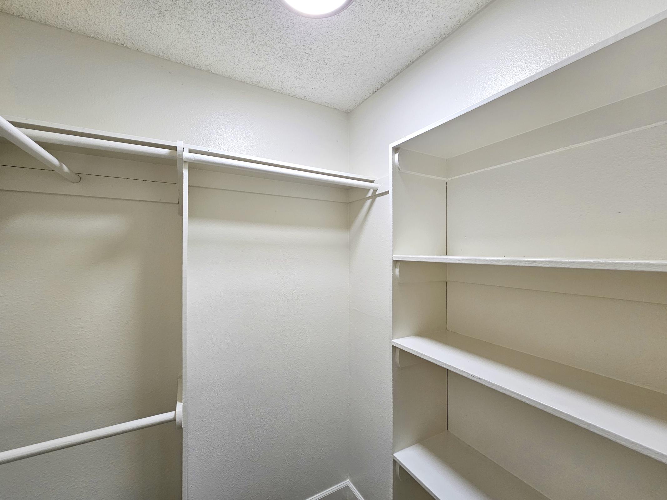 A small, empty closet with white walls. There are two horizontal hanging rods on the left side and multiple shelves on the right, all painted white. The ceiling has a light fixture, providing bright illumination to the space. The overall appearance is clean and minimalistic.