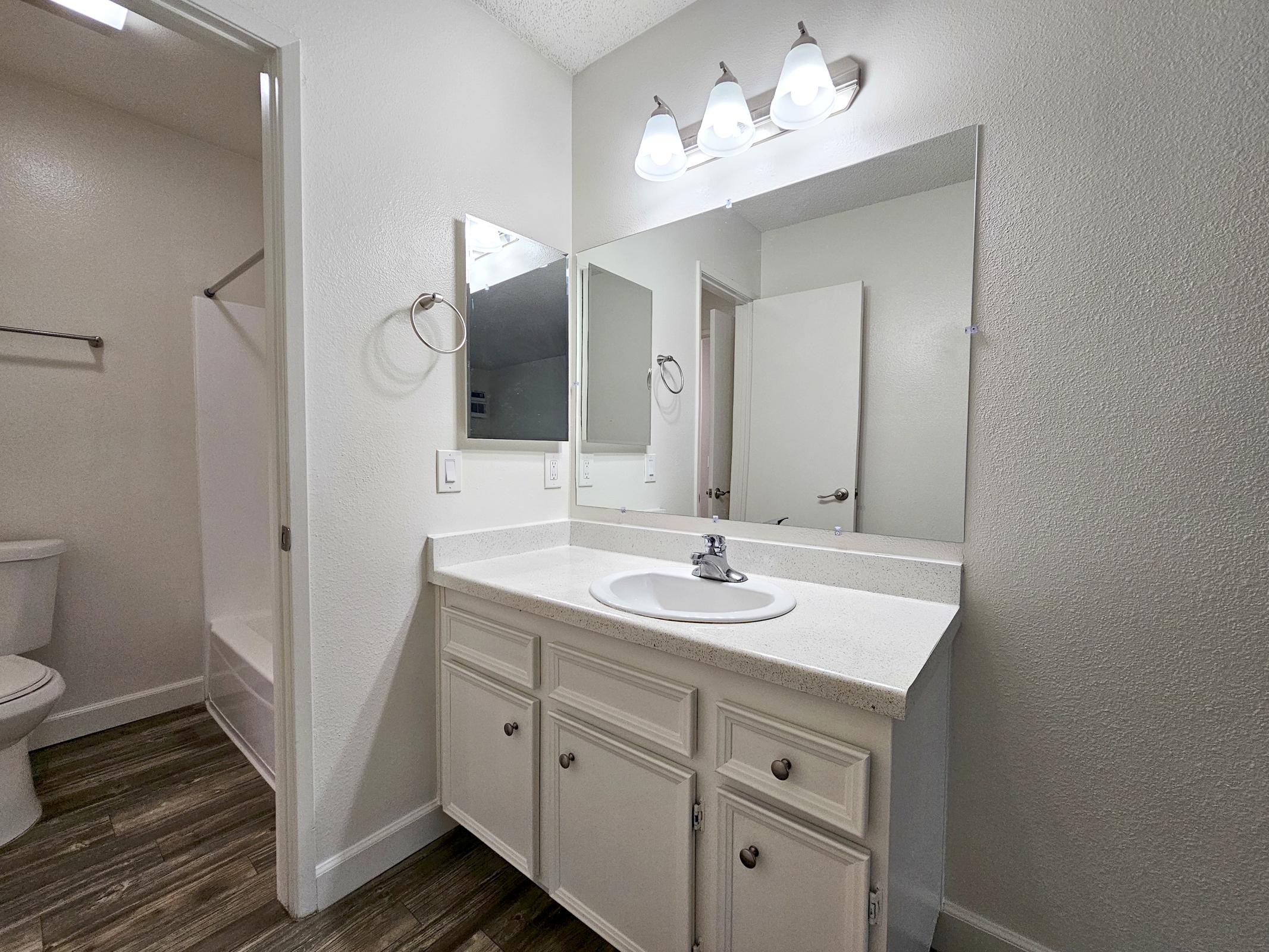 A modern bathroom featuring a clean, white vanity with a sink and polished chrome faucet. Above the vanity, three light fixtures illuminate the space. A large mirror reflects the room. On the left, there is a toilet and shower area, with light-colored walls and wood-like flooring.