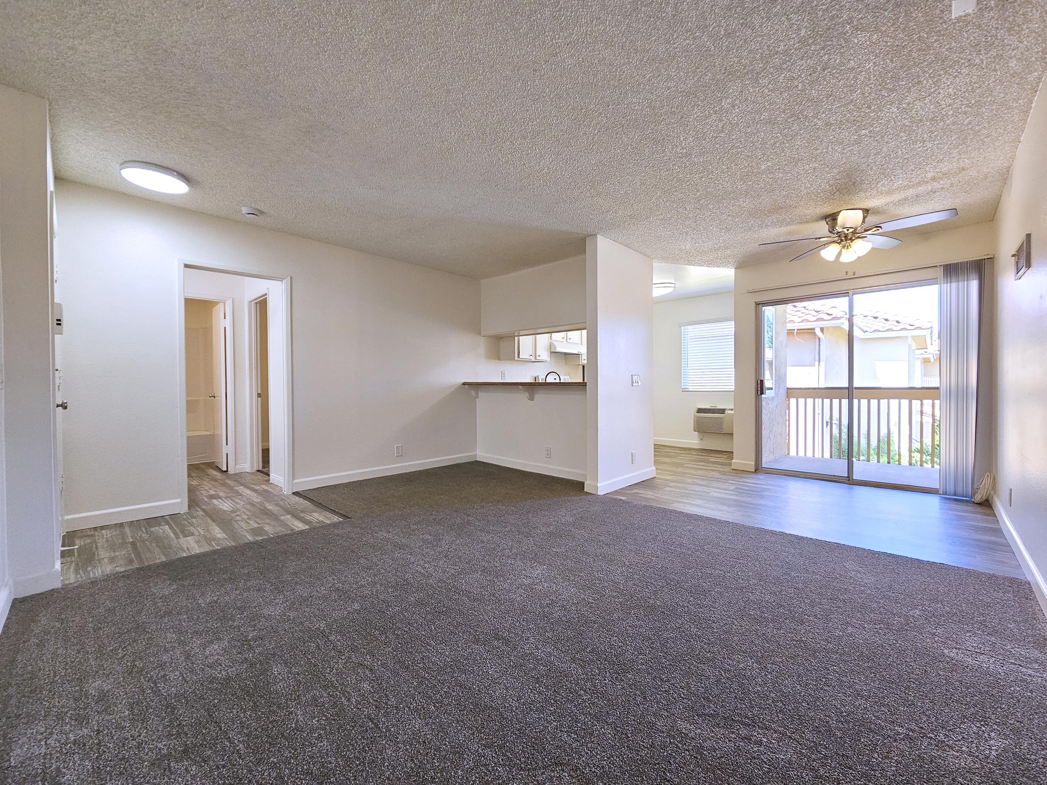 Spacious living area with a light-colored carpet, featuring a ceiling fan and natural light from a sliding glass door that opens to a balcony. The room includes a small kitchen area with a counter, and there is an adjacent hallway leading to another room, all in a neutral color scheme.