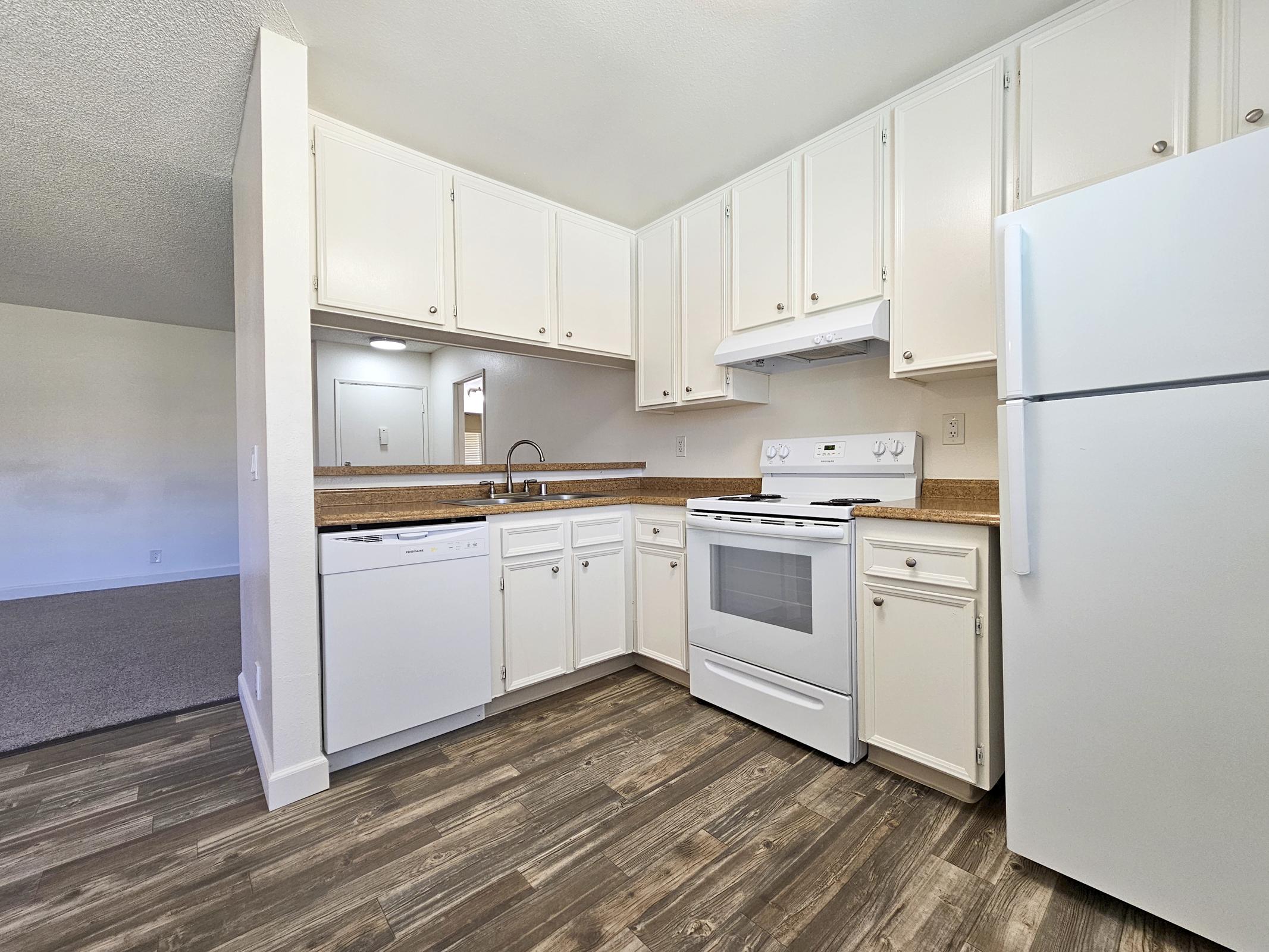Bright, modern kitchen featuring white cabinetry, a white refrigerator, an oven, and a dishwasher. The countertops are a warm brown with a wood-like floor. A spacious layout includes a sink and ample storage, with natural light entering from a nearby window or doorway.