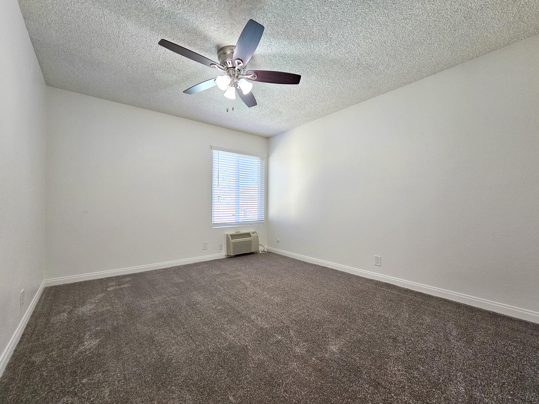 A spacious, empty room with a ceiling fan and light fixtures. The walls are painted white, and there is a window with blinds letting in natural light. The floor is carpeted in a light shade, and there is an air conditioning unit along one wall, creating a bright and airy atmosphere.