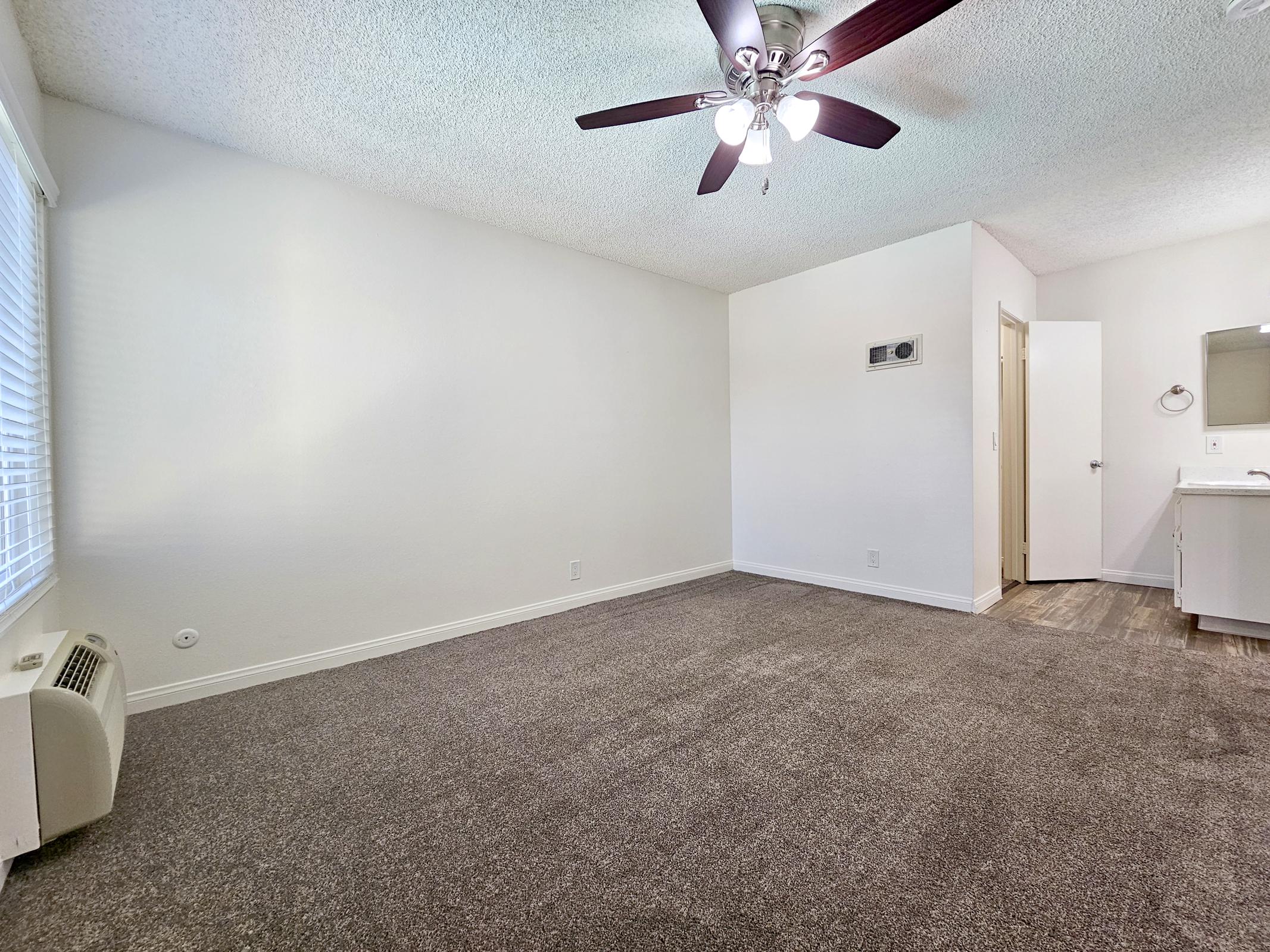 An empty room with carpeted flooring, bright white walls, and a ceiling fan. A wall-mounted air conditioning unit is visible, along with a doorway leading to a bathroom. Natural light shines through a window with blinds, creating a spacious, airy atmosphere. The decor is minimal and modern.
