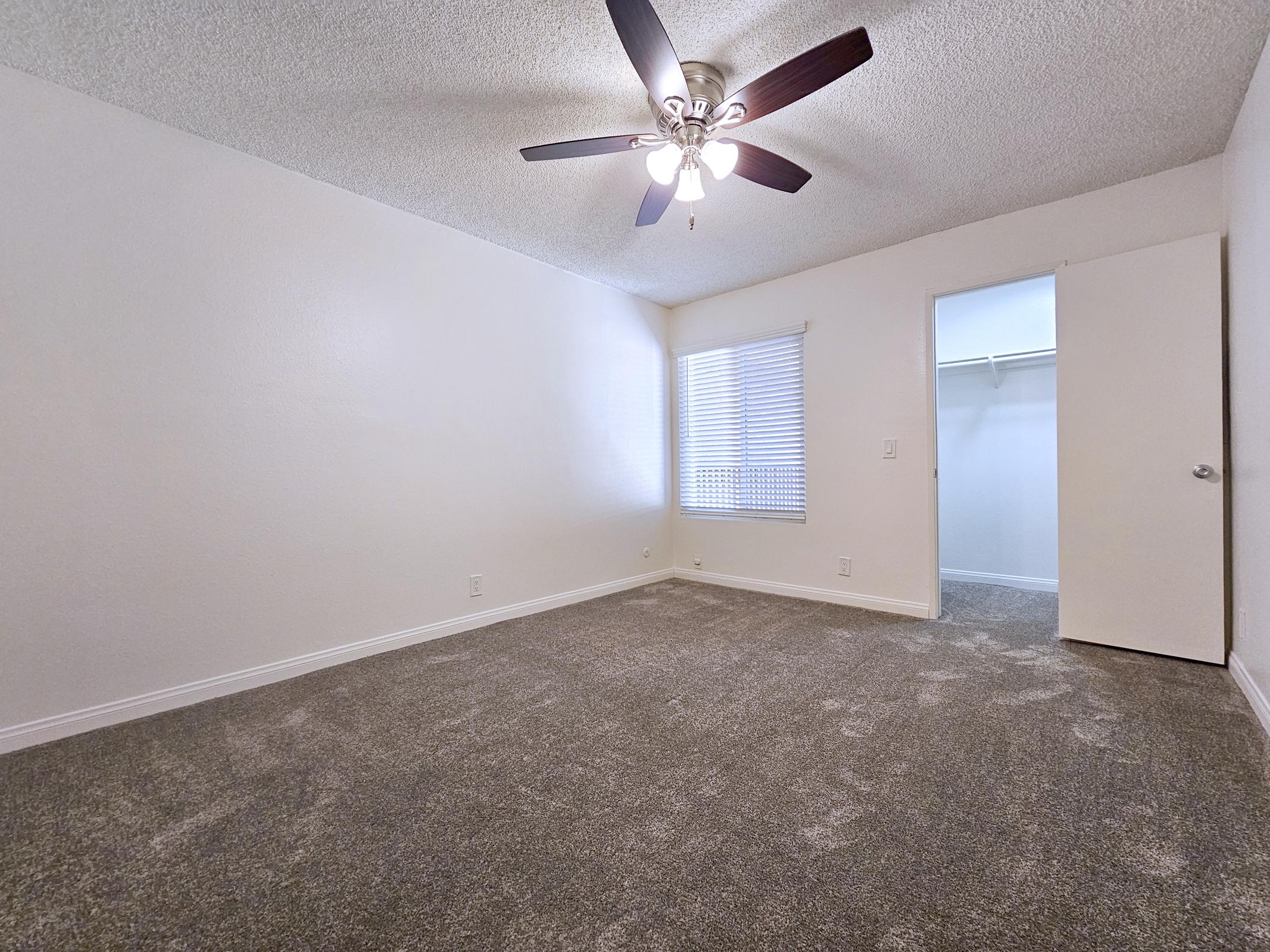 A spacious, well-lit bedroom featuring a ceiling fan, light-colored walls, and plush carpeting. There is a window with blinds allowing natural light, along with a closed closet on one side of the room. The overall ambiance is clean and inviting, ideal for a comfortable living space.