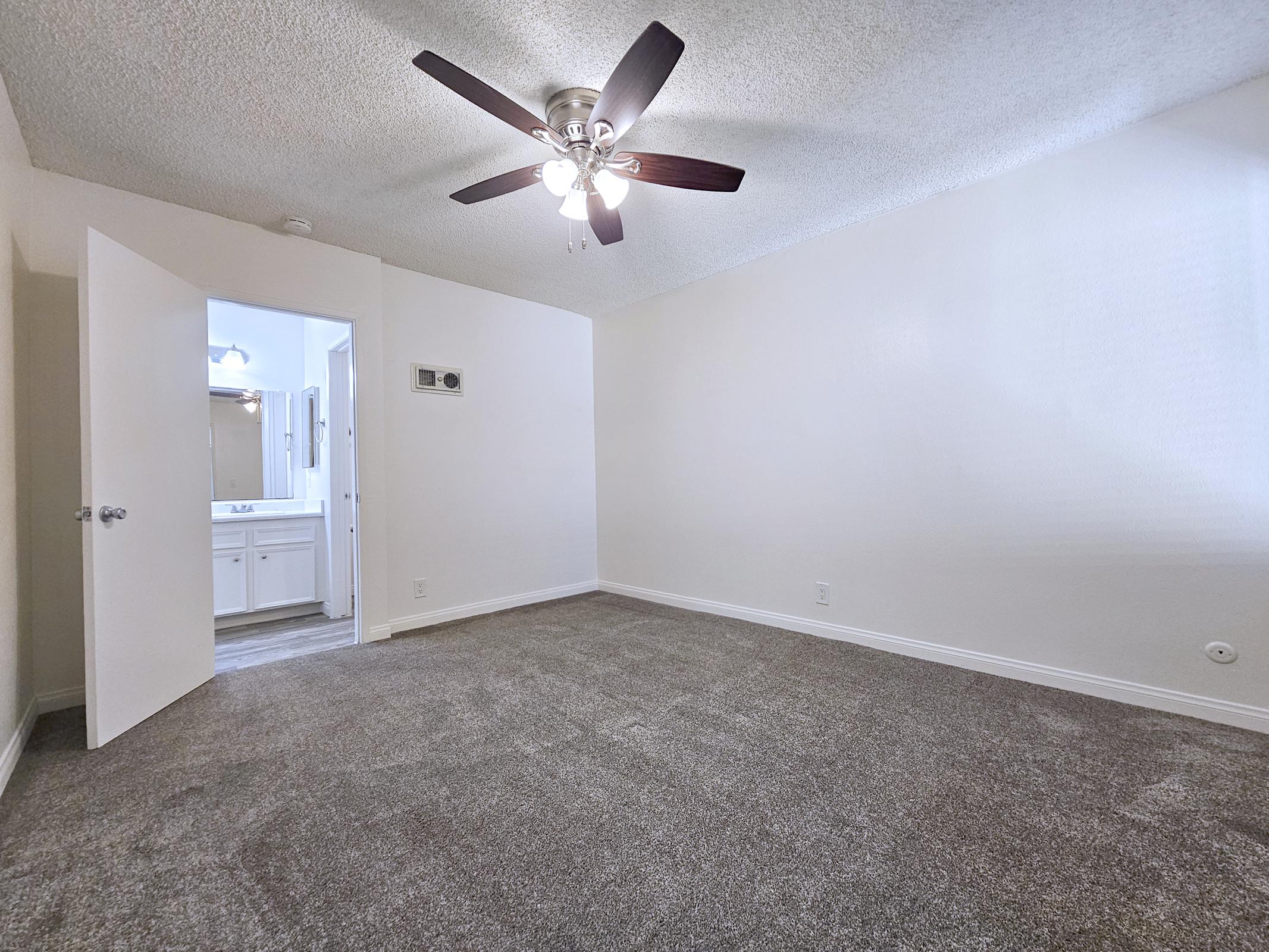 A spacious, well-lit room with a ceiling fan and soft carpet. There is an open door leading to a bathroom, visible in the background. The walls are painted white, creating a clean and fresh atmosphere. The overall ambiance is inviting and suitable for relaxation or personal use.