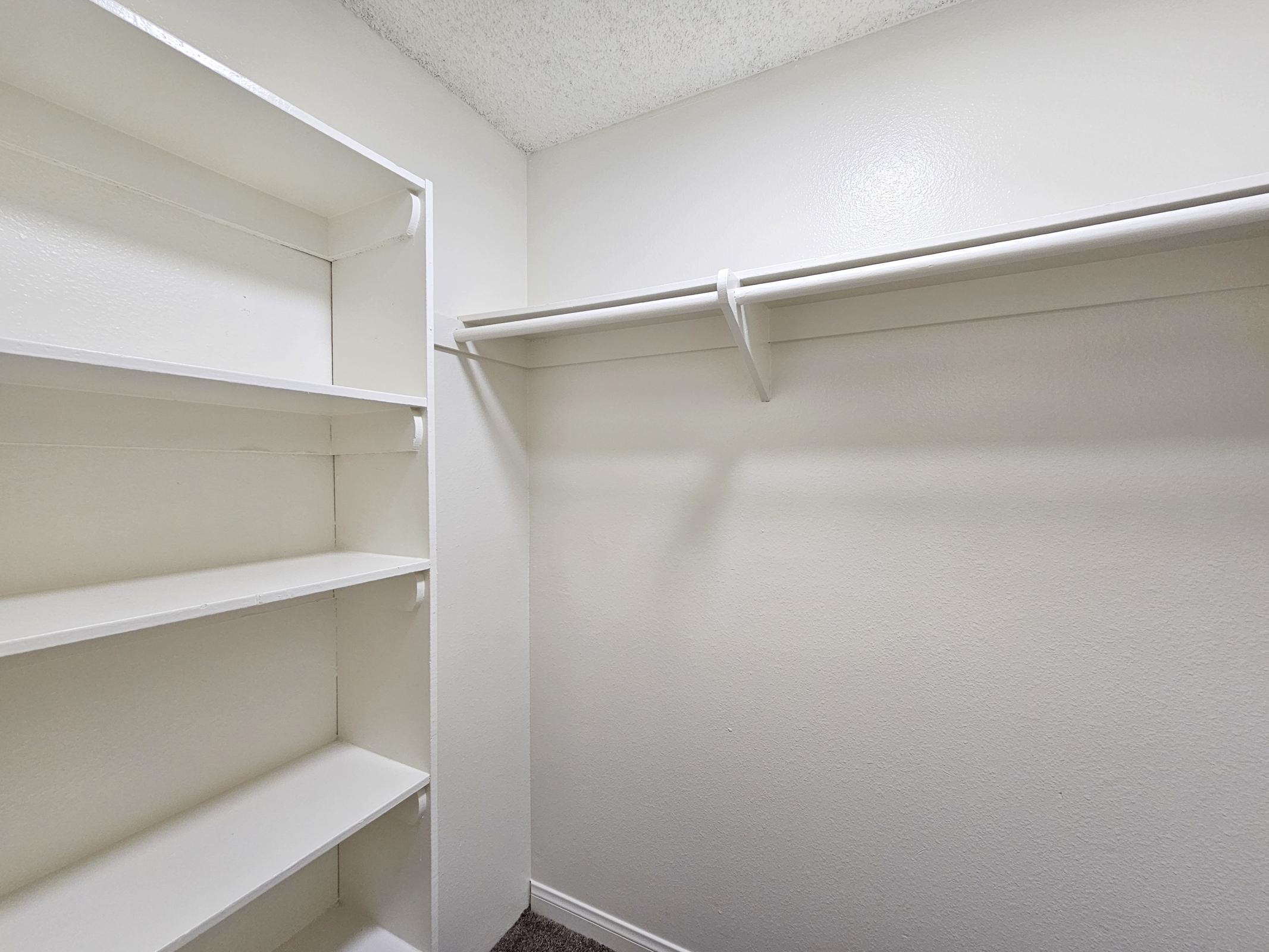 Empty closet with white walls and shelving. The left side features built-in shelves, while the right side has a hanging rod for clothes. The floor is carpeted, and the overall space appears tidy and minimalistic.