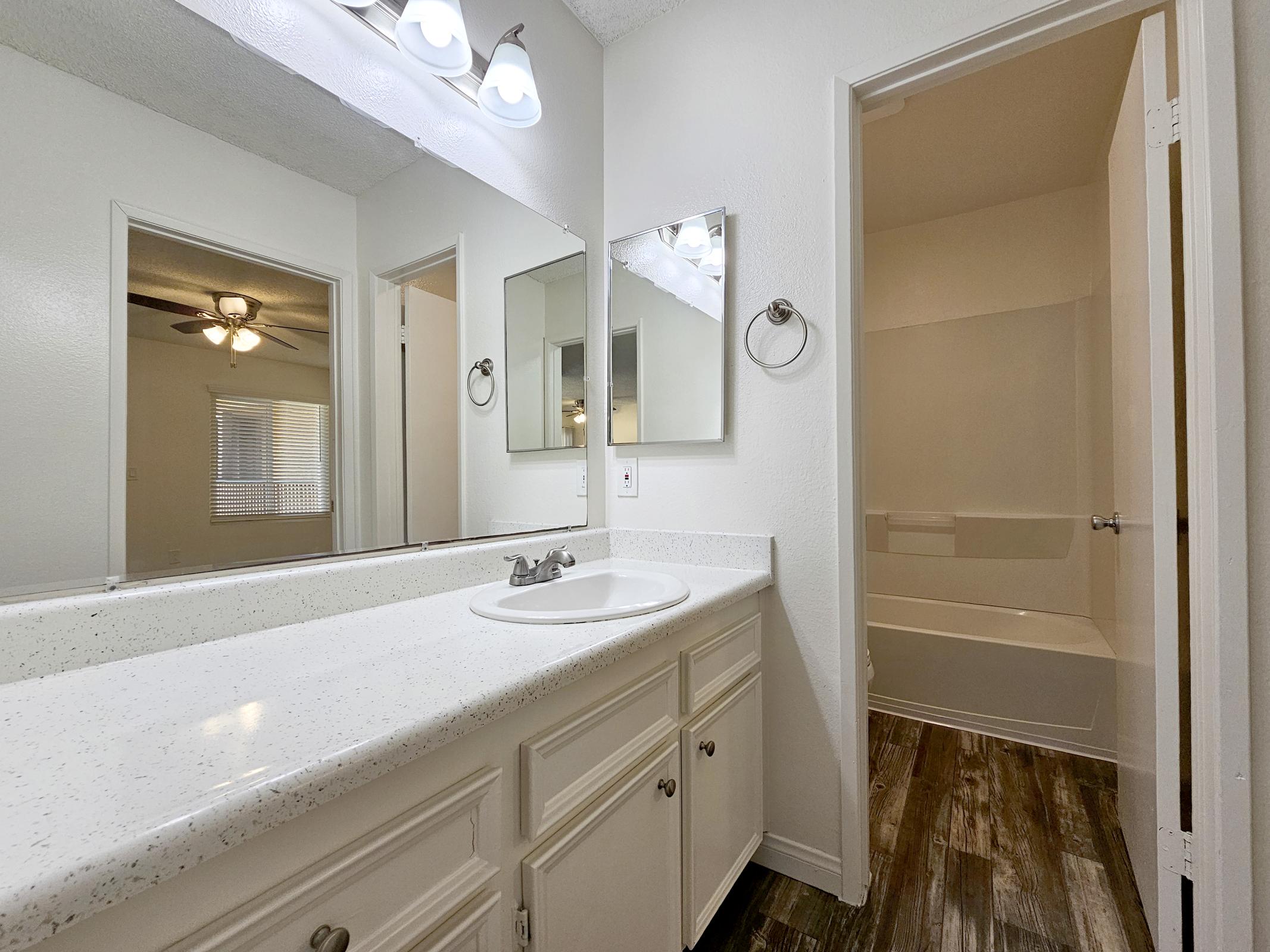A well-lit bathroom featuring a double sink vanity with a mirror, light fixtures above, and a door leading to a tub and shower area. The flooring is a wood-like laminate, and there are large mirrors on the wall, providing an open and airy feel to the space.
