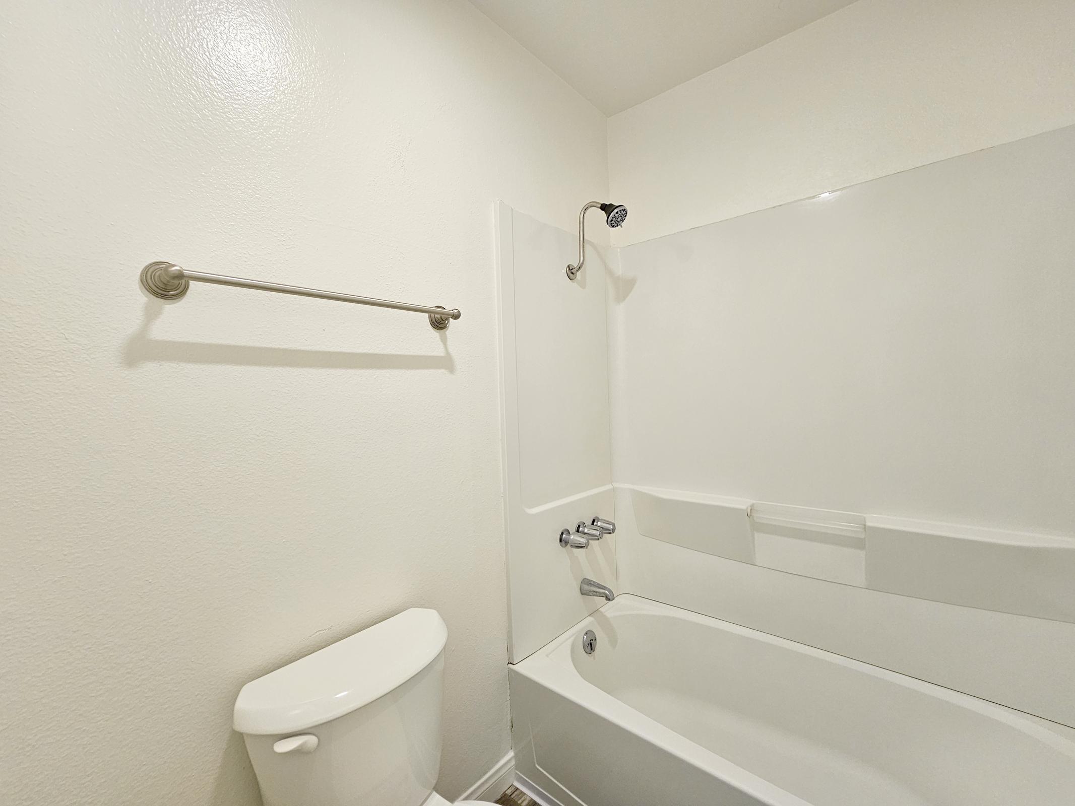 A clean and minimalistic bathroom featuring a white bathtub with a showerhead attached, a toilet, and a towel bar on the wall. The walls are painted in a light color, and the space is bright and uncluttered, showcasing a simple and modern design.
