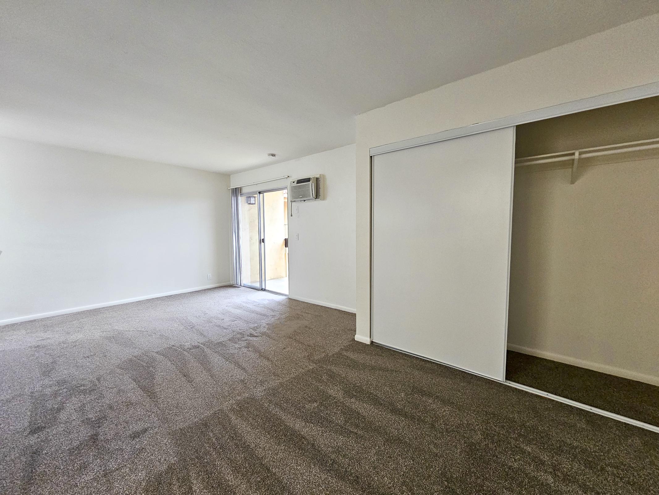Spacious interior of a room featuring light-colored walls, carpeted flooring, and a large sliding door leading to an outdoor area. There's a closet with sliding doors on the right. The room is well-lit, showcasing a clean and modern design. An air conditioning unit is visible on the wall.