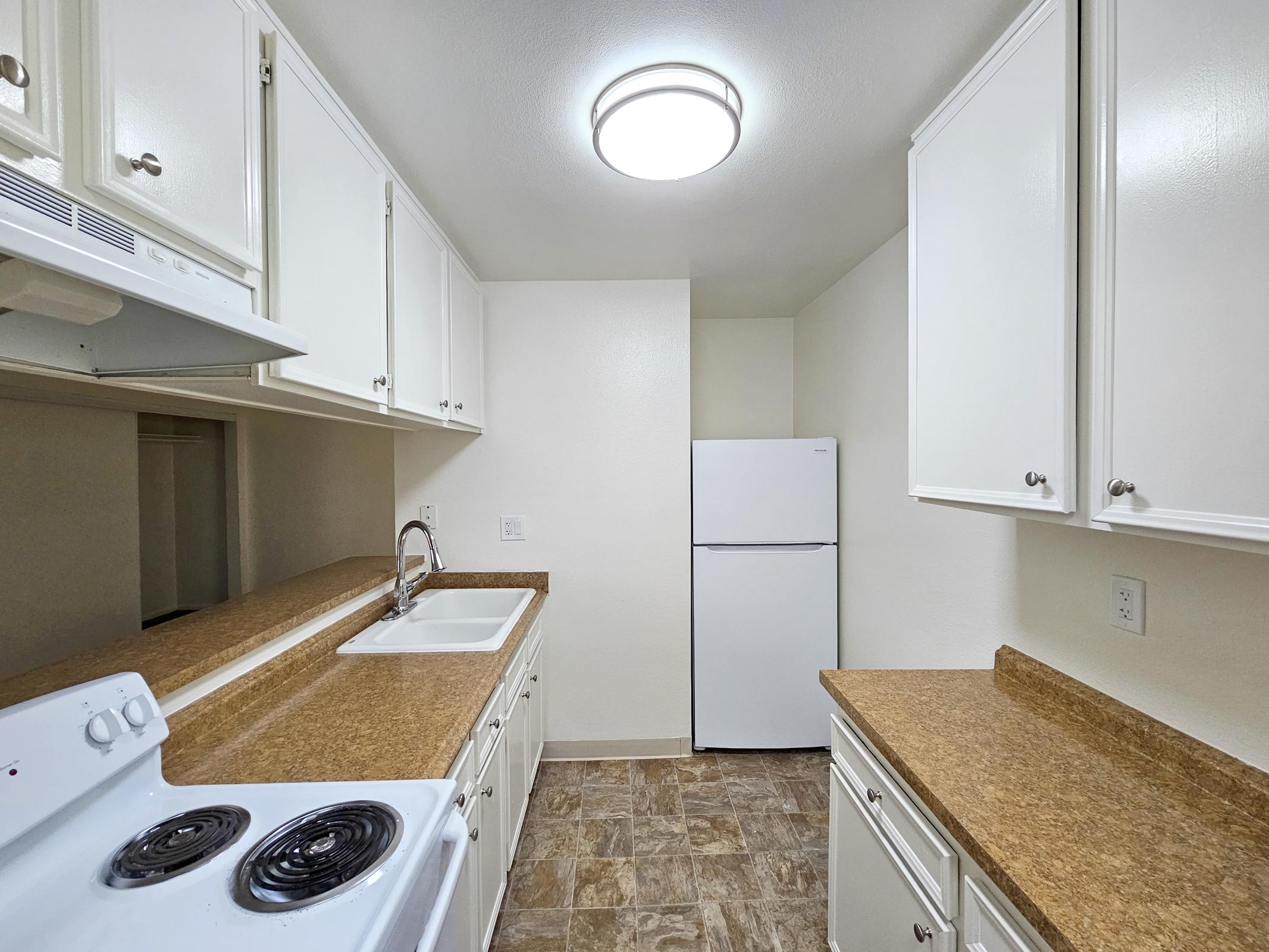 A clean, modern kitchen with white cabinets, a sink, a stove, and a refrigerator. The countertop is a light brown material, and the flooring is a patterned tile. A circular ceiling light illuminates the space, giving it a bright and airy feel. The layout is functional, with ample storage and workspace.