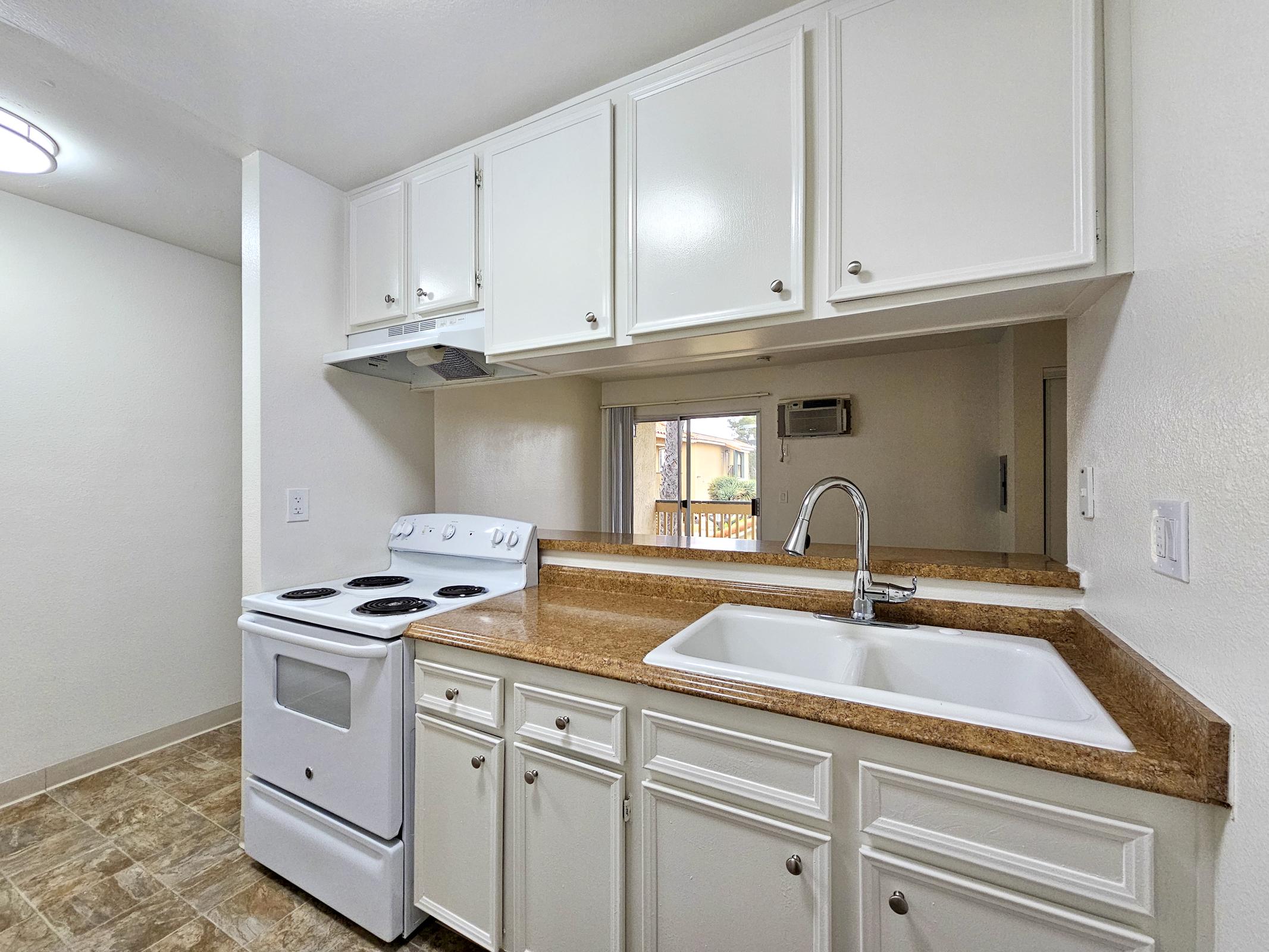 Bright and clean kitchen featuring white cabinets, a stainless steel sink, and an electric stove. The countertop is a light brown granite, and the floor has beige tiles. A window offers natural light, illuminating the space, which is designed with a modern and inviting aesthetic.