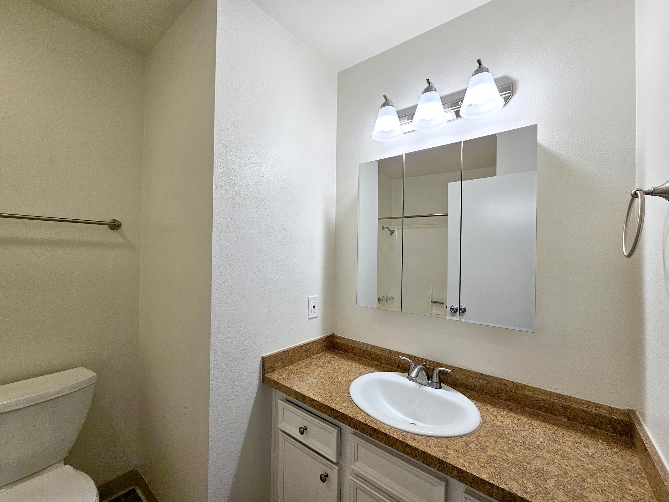 A clean and bright bathroom featuring a white sink with a simple chrome faucet, a large mirror above the sink, and a light fixture with three bulbs. The countertop is made of brown stone. A toilet is visible in the corner, with a towel bar mounted on the wall, and a shower area can be seen through a glass door.