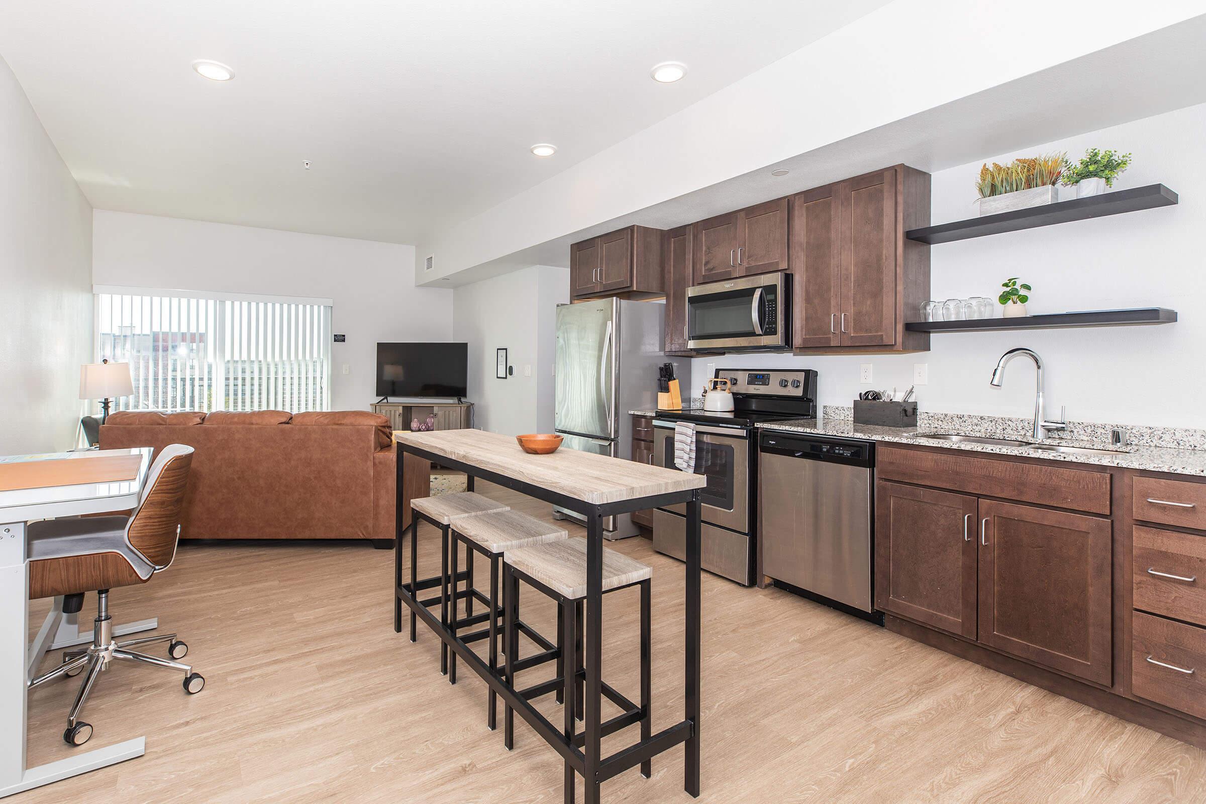 Modern kitchen and dining area featuring wooden cabinetry, stainless steel appliances, a microwave, a kitchen cart with cookware, a round dining table with four chairs, and a cozy living space with a brown sofa and large window. The decor is minimalist with neutral tones and touches of contemporary design.