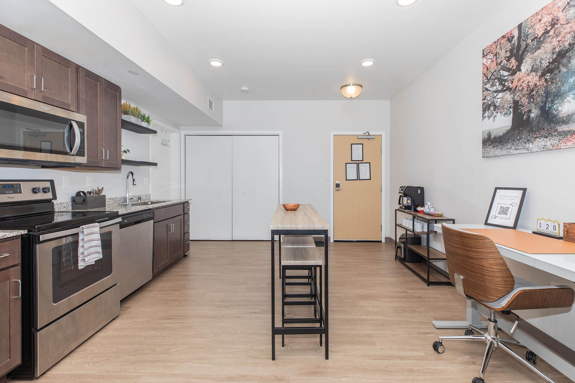 A modern kitchen and dining area featuring a round wooden table with chairs, an open layout, stainless steel appliances, and a countertop. The space has light wood flooring, minimalistic decor, and large walls with a simple color scheme, creating a bright and inviting atmosphere.