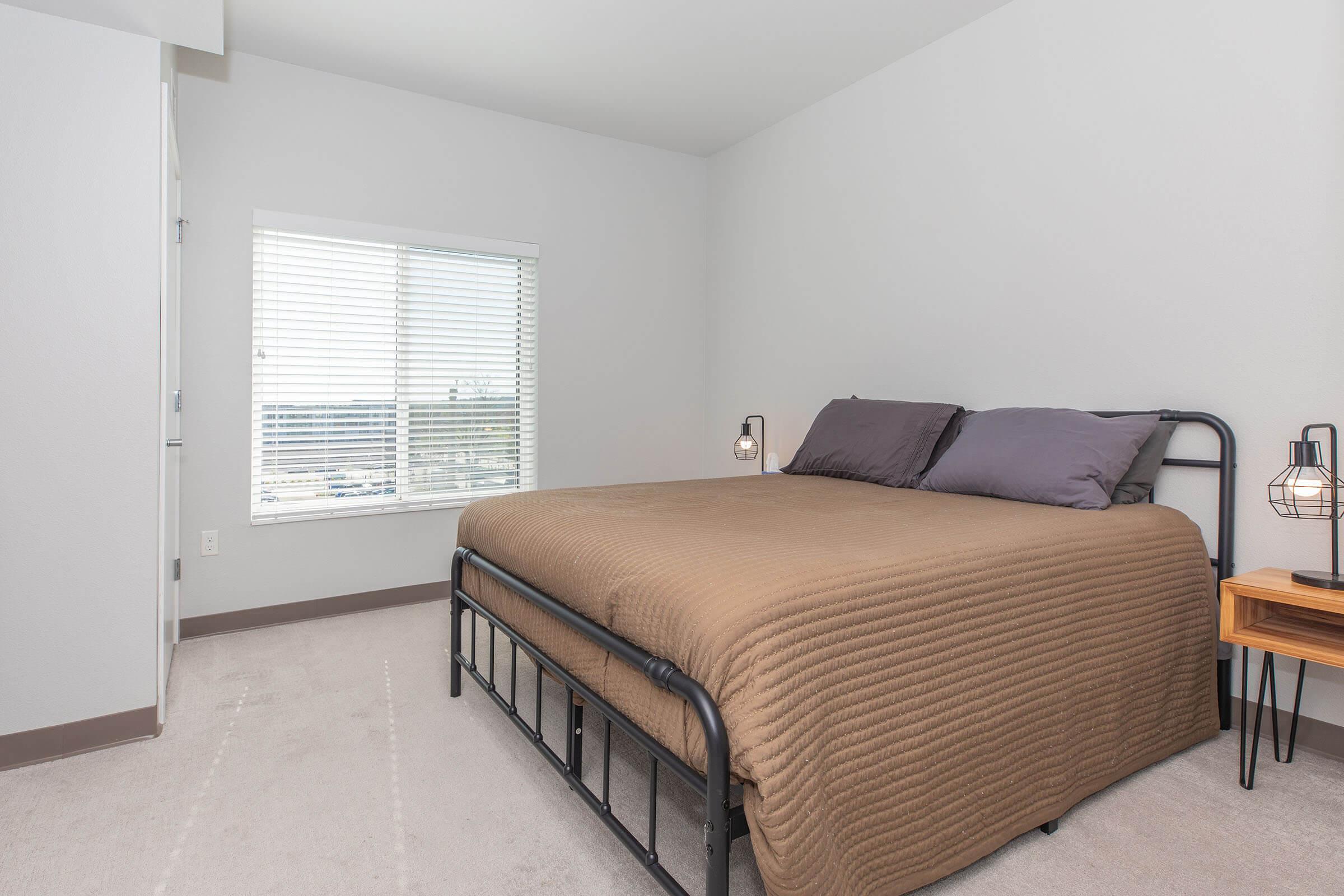 A cozy bedroom featuring a metal bed frame with a neatly made bed, a small bedside table with a lamp, and a large mirror. Natural light streams in through a window with blinds, illuminating the light-colored walls and wooden floor, creating a warm and inviting atmosphere. Two framed artworks are hanging on the wall.