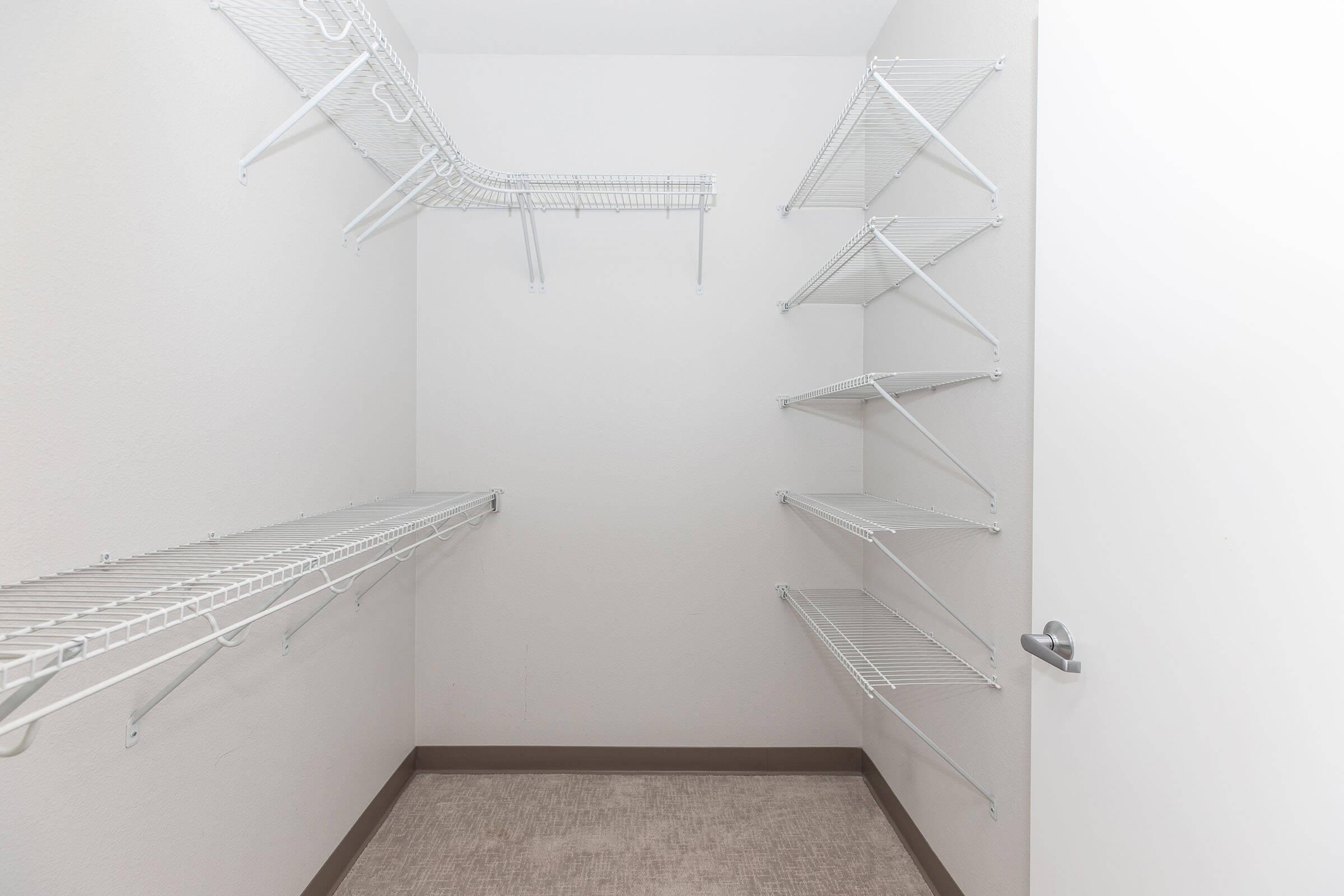 A clean, modern bathroom featuring a white toilet, a sink with a granite countertop, and a large mirror. Handrails are installed for accessibility. There is a shelf with neatly folded gray towels and a trash can nearby. The floor is tiled, and the walls are painted light gray.