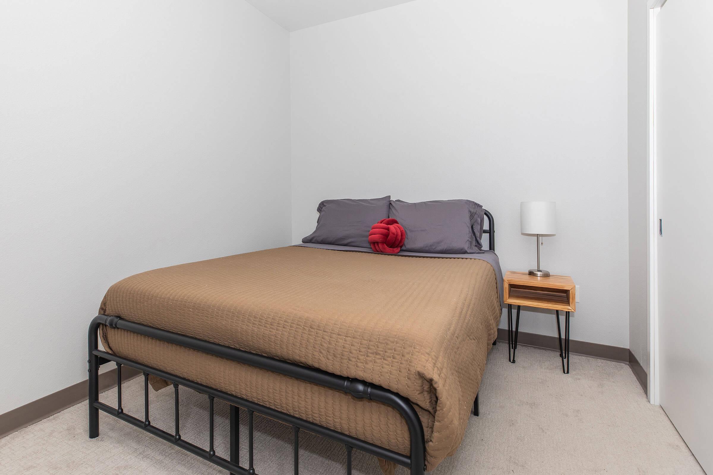 A cozy bedroom featuring a soft brown quilt on a twin-sized bed with gray pillows. Next to the bed is a small wooden nightstand with a lamp. The walls are light gray, and there are two simple wall decorations above the bed, adding a modern touch to the room's design.