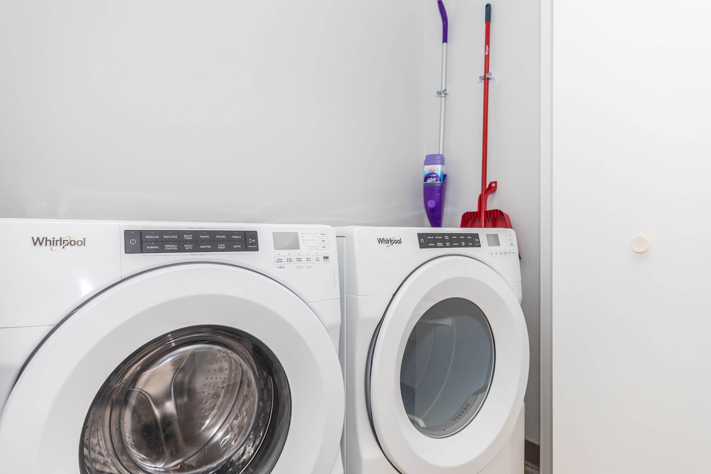 A set of white Whirlpool washer and dryer machines side by side in a laundry room. The wall behind them is light-colored, and there are cleaning tools, including a purple vacuum and a red broom, hanging on the wall.