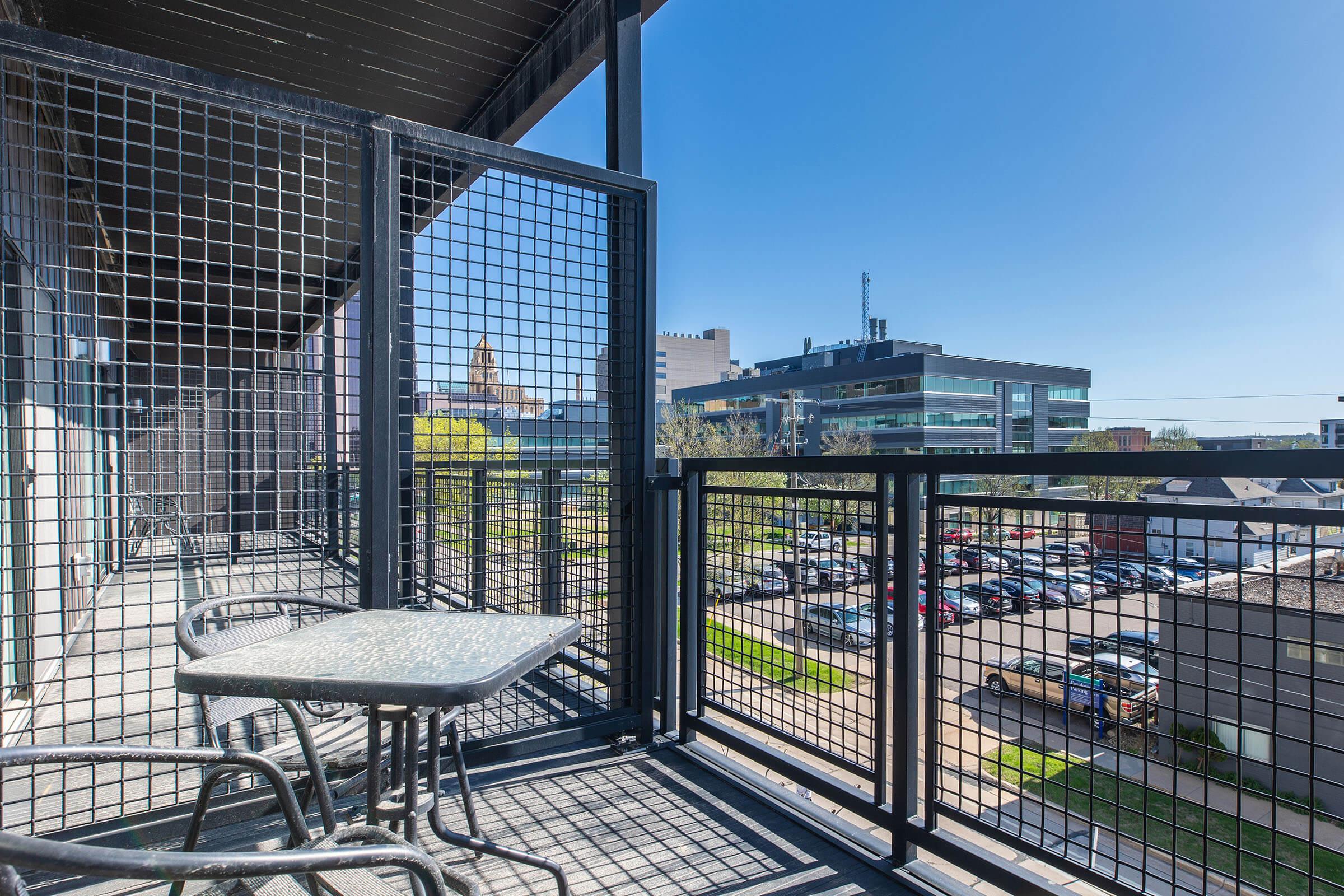 A small balcony featuring a round table and two chairs, with a clear blue sky above. In the background, a modern building and a parking lot can be seen, highlighted by greenery in the surrounding area. The balcony has a black metal railing and wooden paneling on the walls.