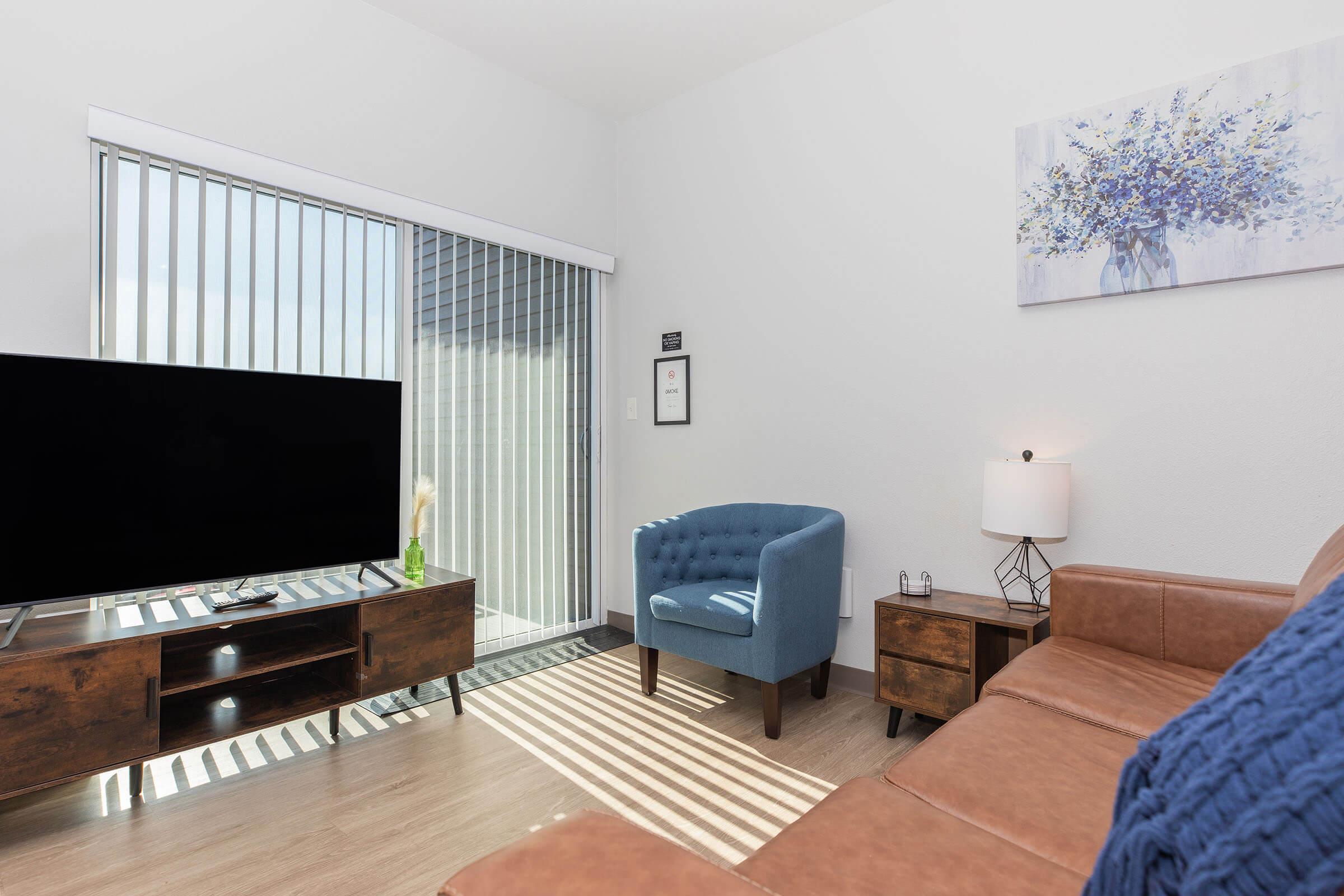 A cozy living room featuring a brown leather couch, a blue upholstered armchair, and a wooden TV stand with a large television. Natural light streams in through vertical blinds, highlighting a minimalist decor with a floral painting on the wall and a small lamp on a side table.