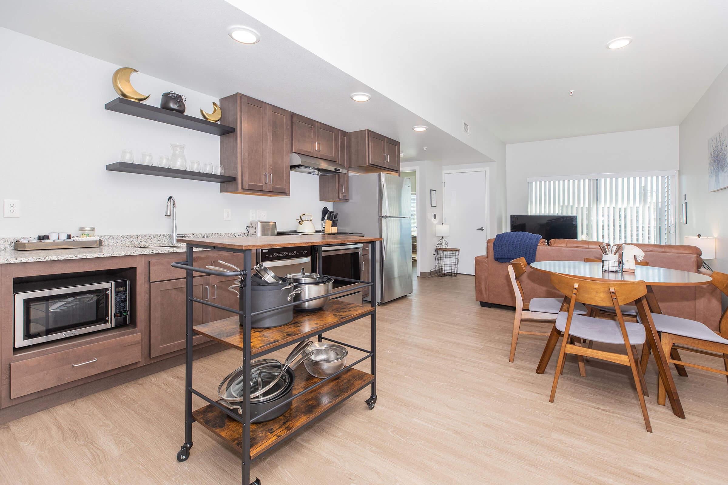 Modern open-concept kitchen and dining area featuring wooden cabinets, a granite countertop, stainless steel appliances, and a dining table with four chairs. A microwave and a cart with cookware are visible. The space is well-lit with natural light coming from nearby windows.