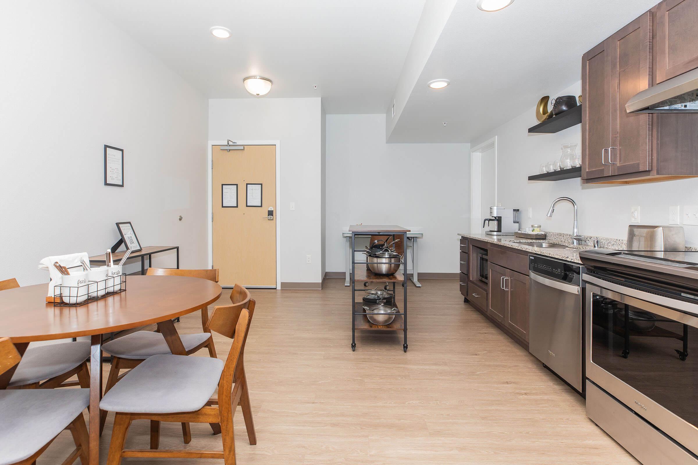 Modern kitchen and dining area featuring a round wooden table with chairs, sleek cabinetry, stainless steel appliances, a countertop with cooking utensils, and a door leading to another room. The space is well-lit with a neutral color scheme and minimalist decor.