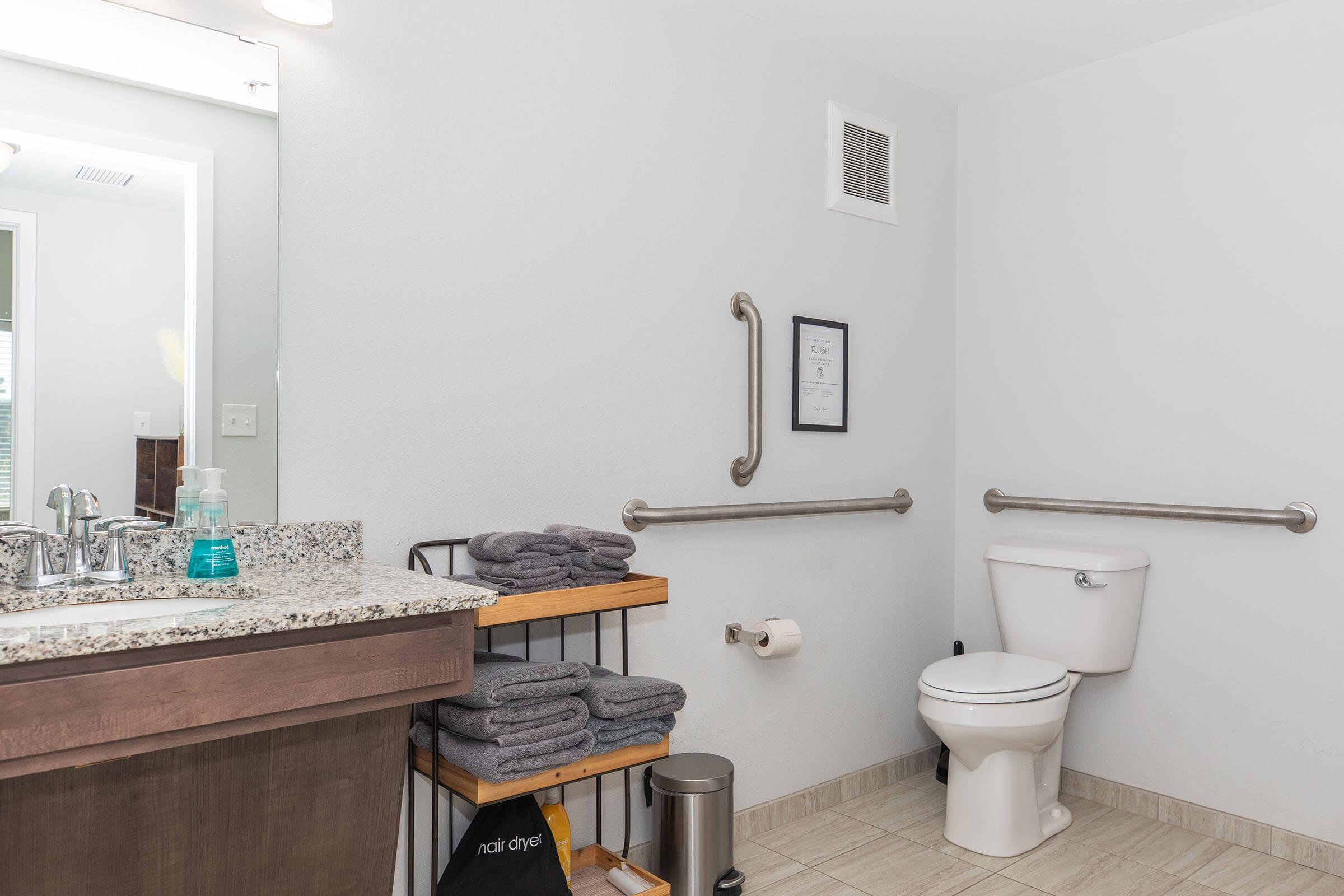 A clean bathroom with a granite countertop sink, a mirror above it, and a roll holder. There are neatly stacked towels on a wooden shelf and a trash can nearby. Grab bars are installed next to the toilet for accessibility, and the space features neutral-colored walls and tiled flooring.
