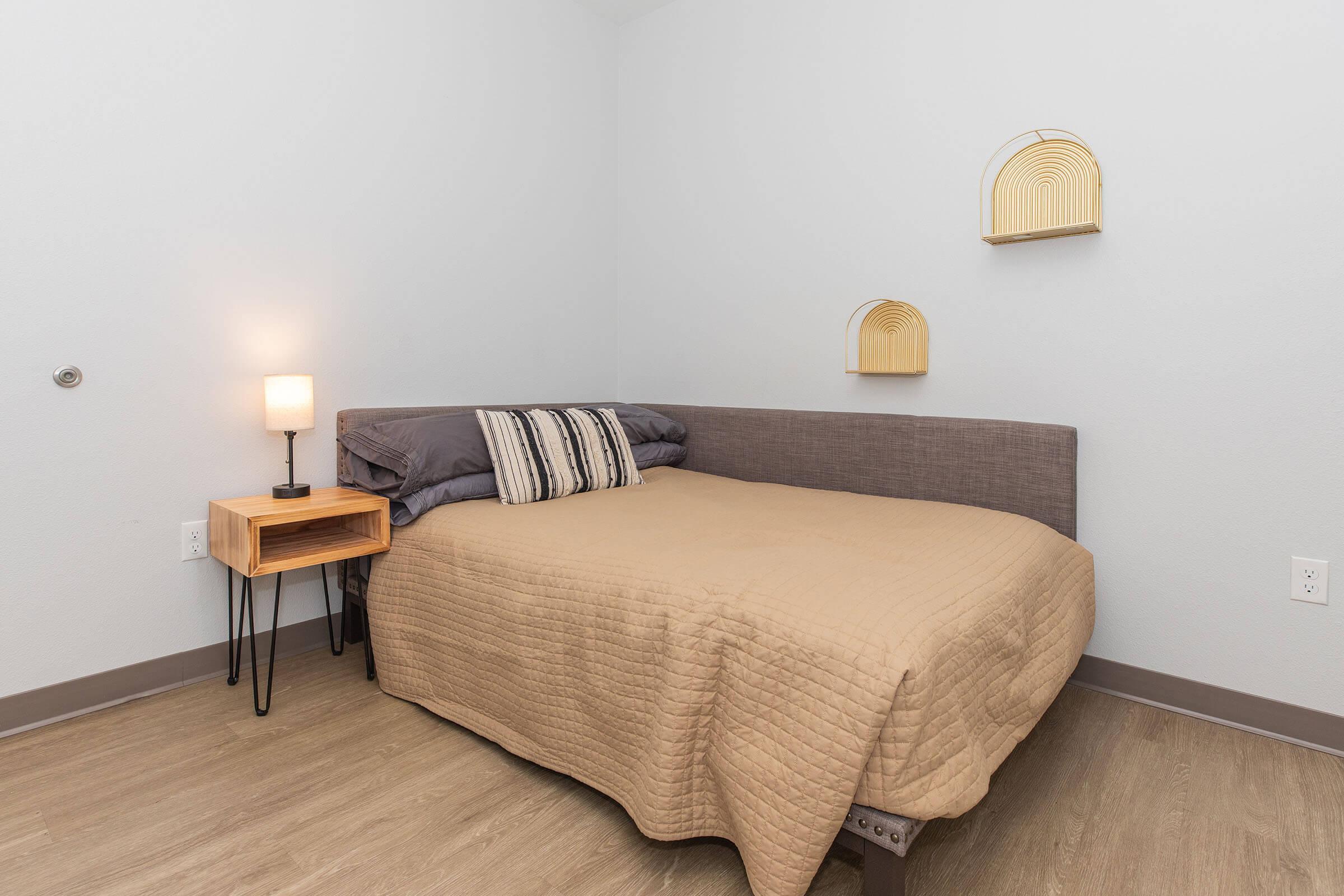 Cozy bedroom featuring a single bed with a beige quilt, accompanied by a small nightstand with a lamp. The walls are light gray, and there are decorative wall shelves with arch-shaped items. The flooring is a warm wood tone, creating a welcoming atmosphere.